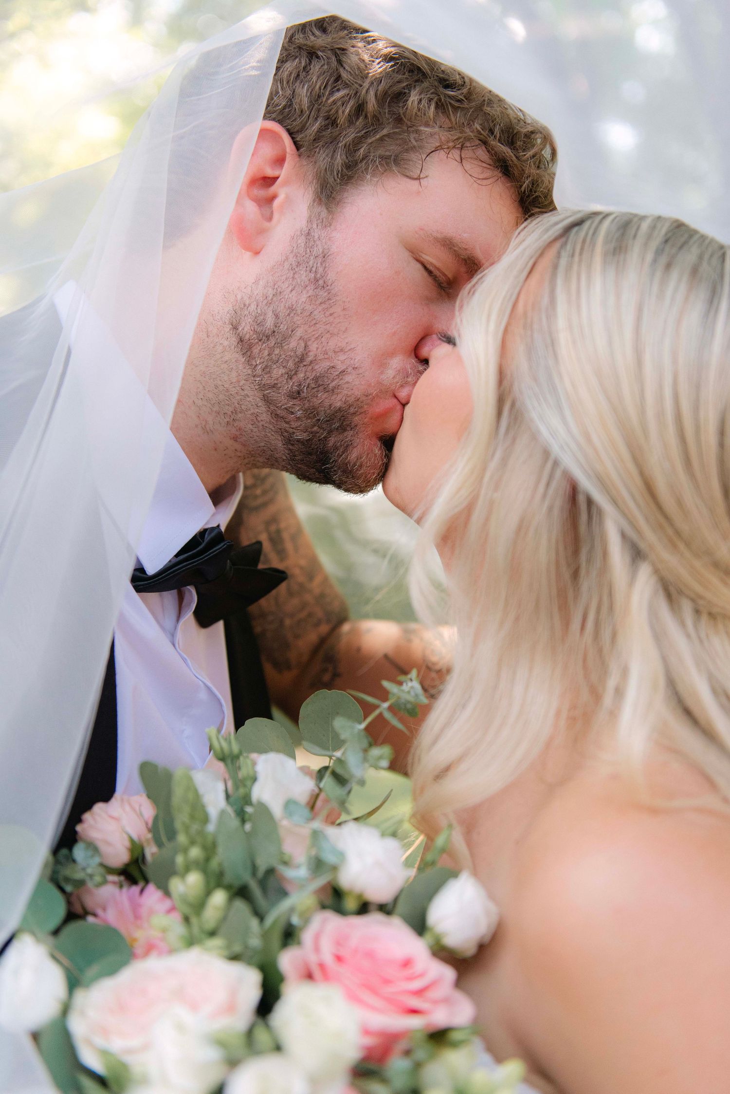 Intimate wedding portrait showing a tender kiss among soft pink and white flowers.