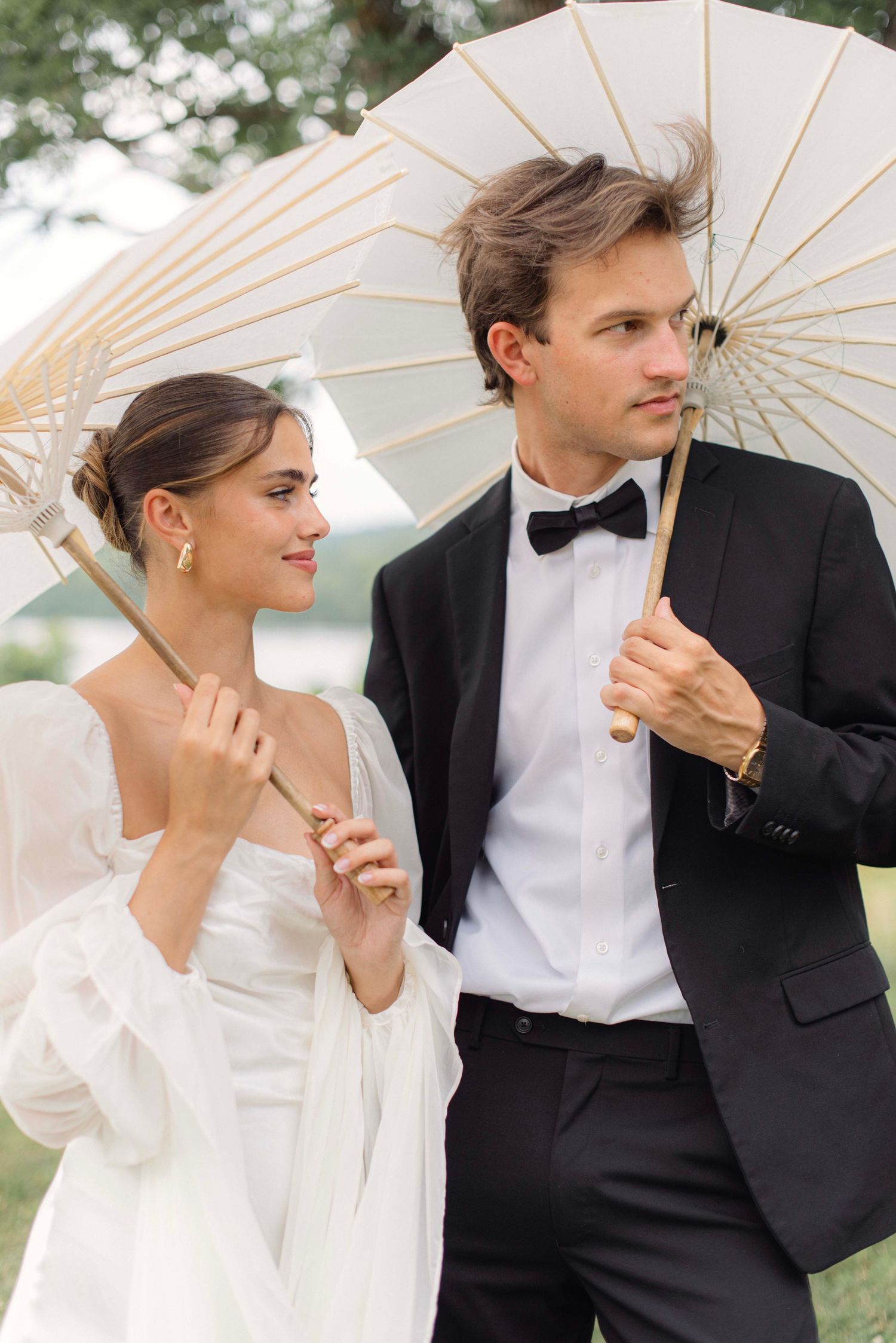 Elegant couple in formal wedding attire posing with white parasols against a scenic outdoor backdrop.