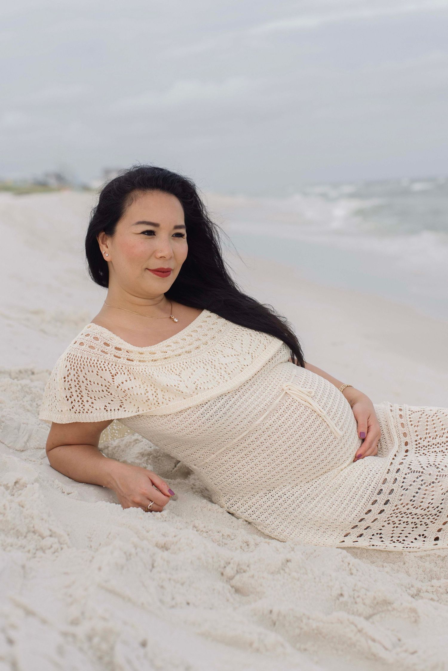 A woman in a cream crochet dress poses on a beach with waves in the background.