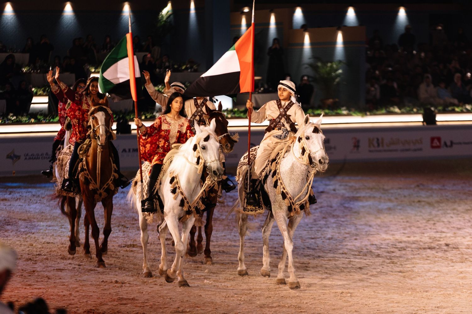Equestrian performers riding in formation during a nighttime show on an illuminated arena stage.