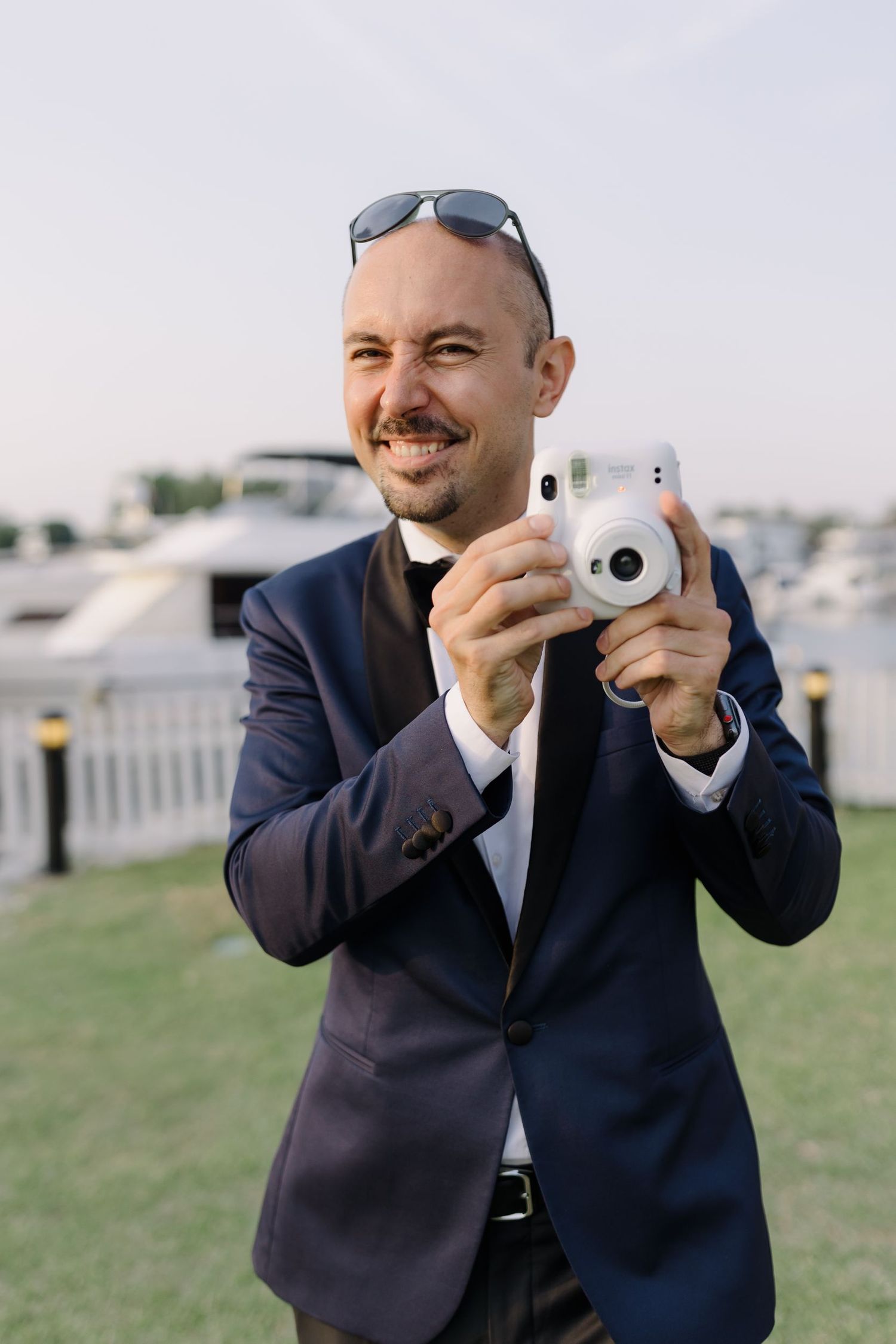 Wedding photographer in navy suit holding an instant camera outdoors.