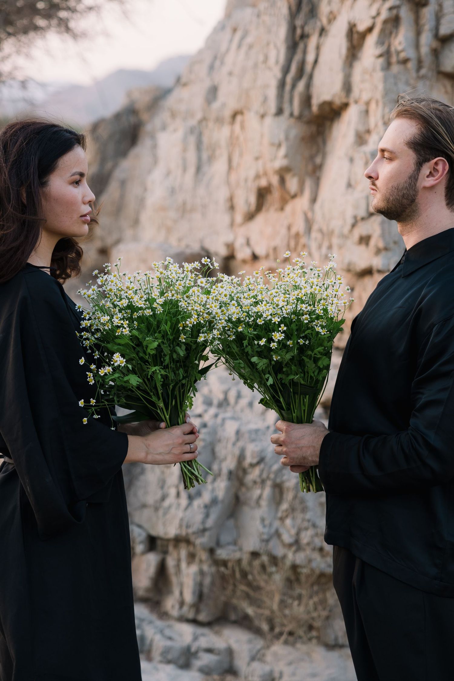 Two people holding white flower bouquets while facing each other against rocky cliffs.