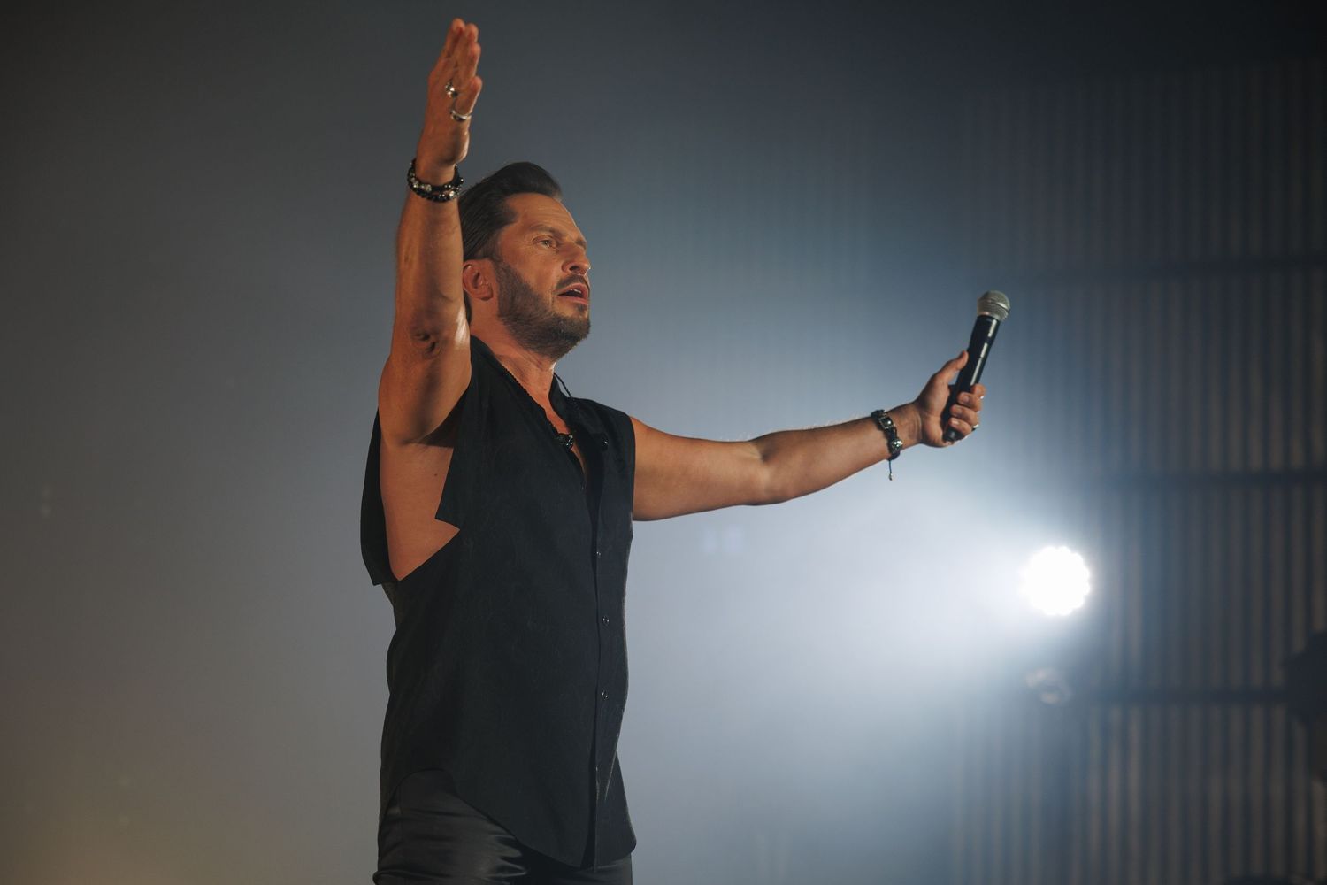 Performer raises arm while holding microphone in dramatic spotlight on stage.