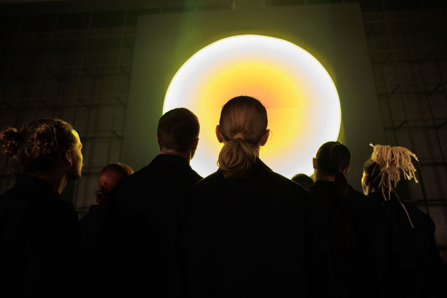 Large glowing yellow orb installation with crowd silhouettes in dark exhibition space.