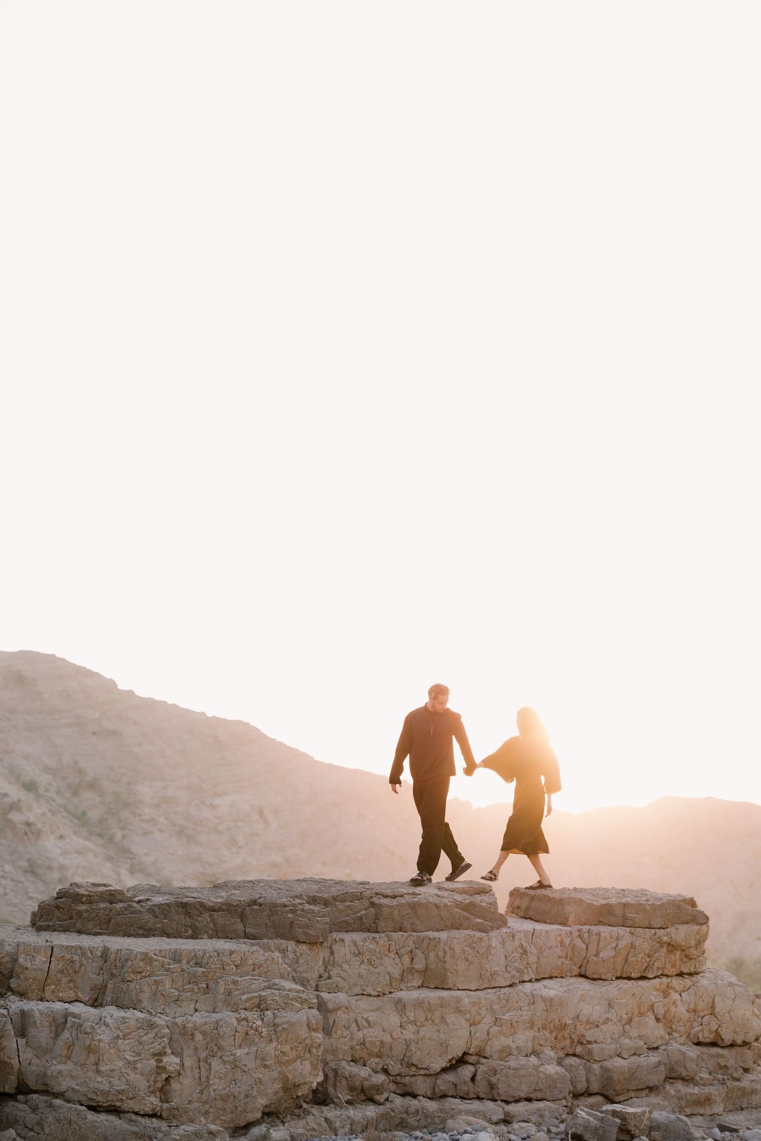 Two silhouettes walk along a rocky cliff edge at sunset.