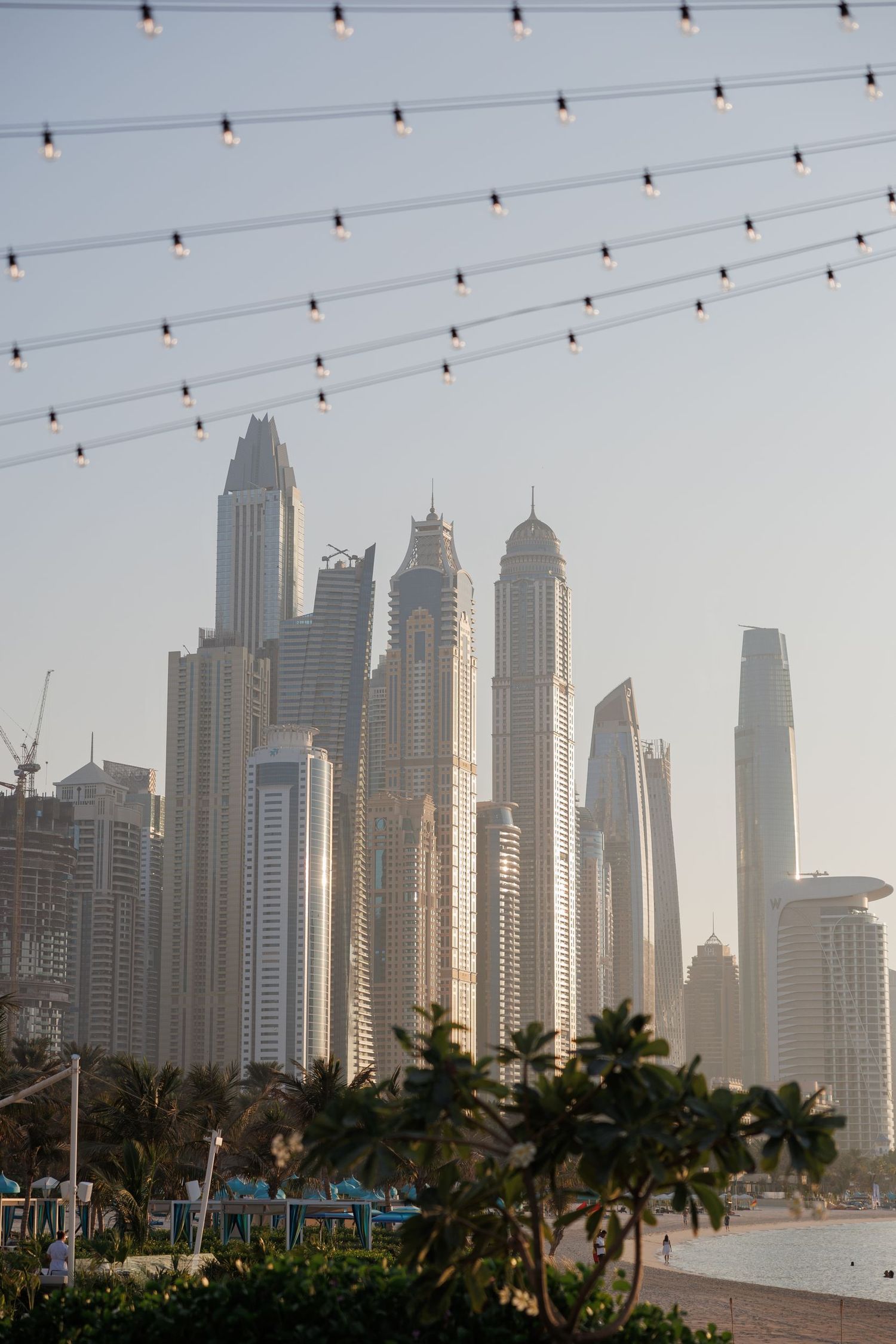 String lights hang above palm trees with Dubai's iconic skyscrapers visible in the misty background.
