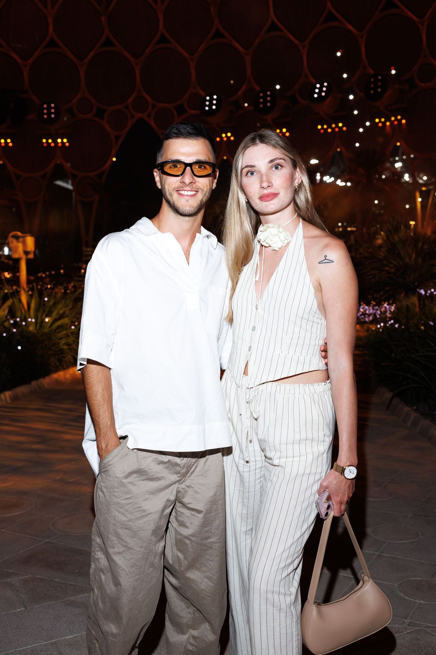 Two people in coordinated white and beige outfits pose together at night.