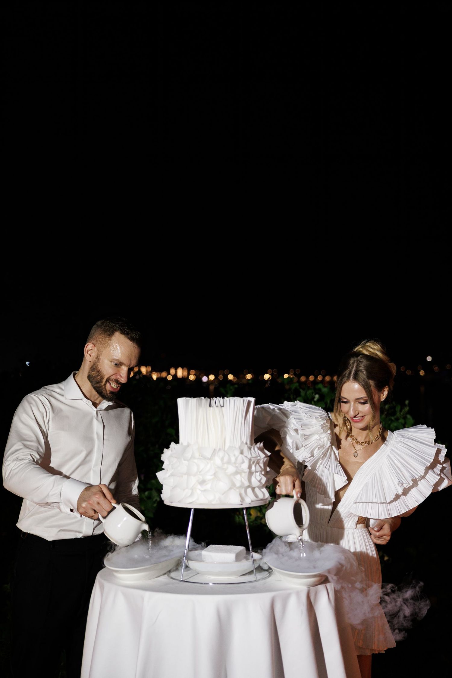 A couple cutting their white tiered wedding cake together at an outdoor evening reception.