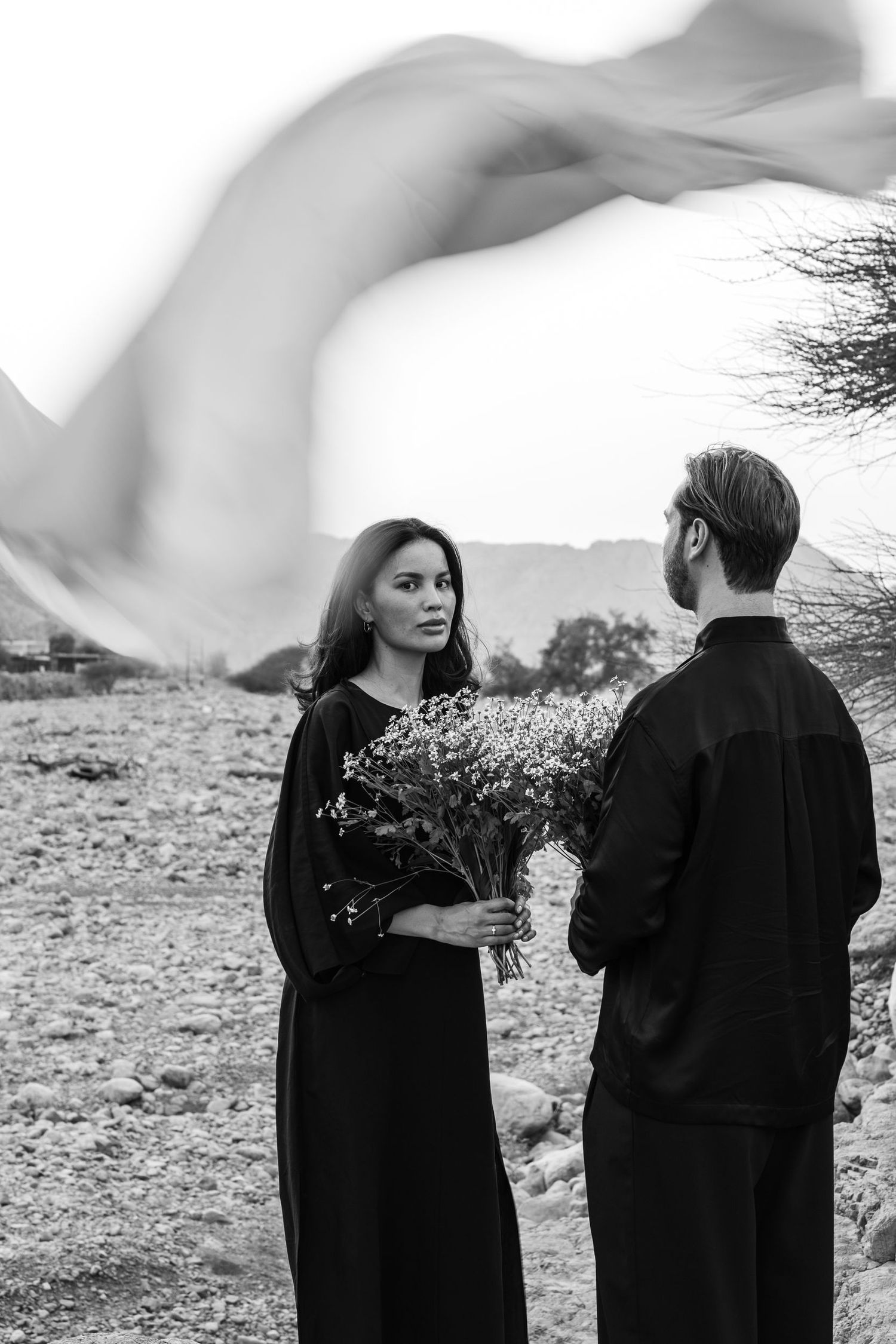 Two figures in black clothing stand against a rocky desert landscape with dramatic flowing fabric.