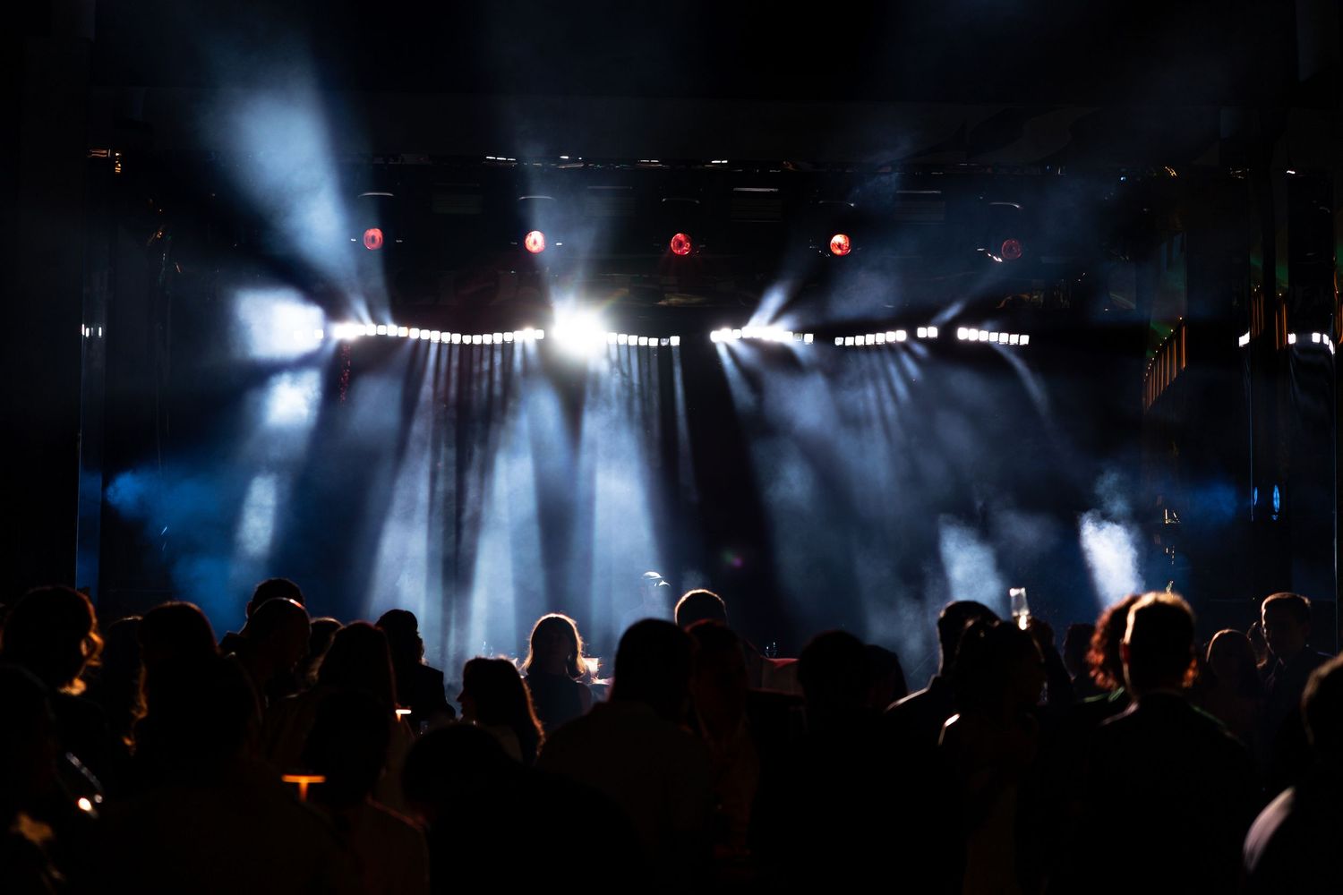 Concert stage illuminated by dramatic white spotlights with silhouetted crowd in dark venue.