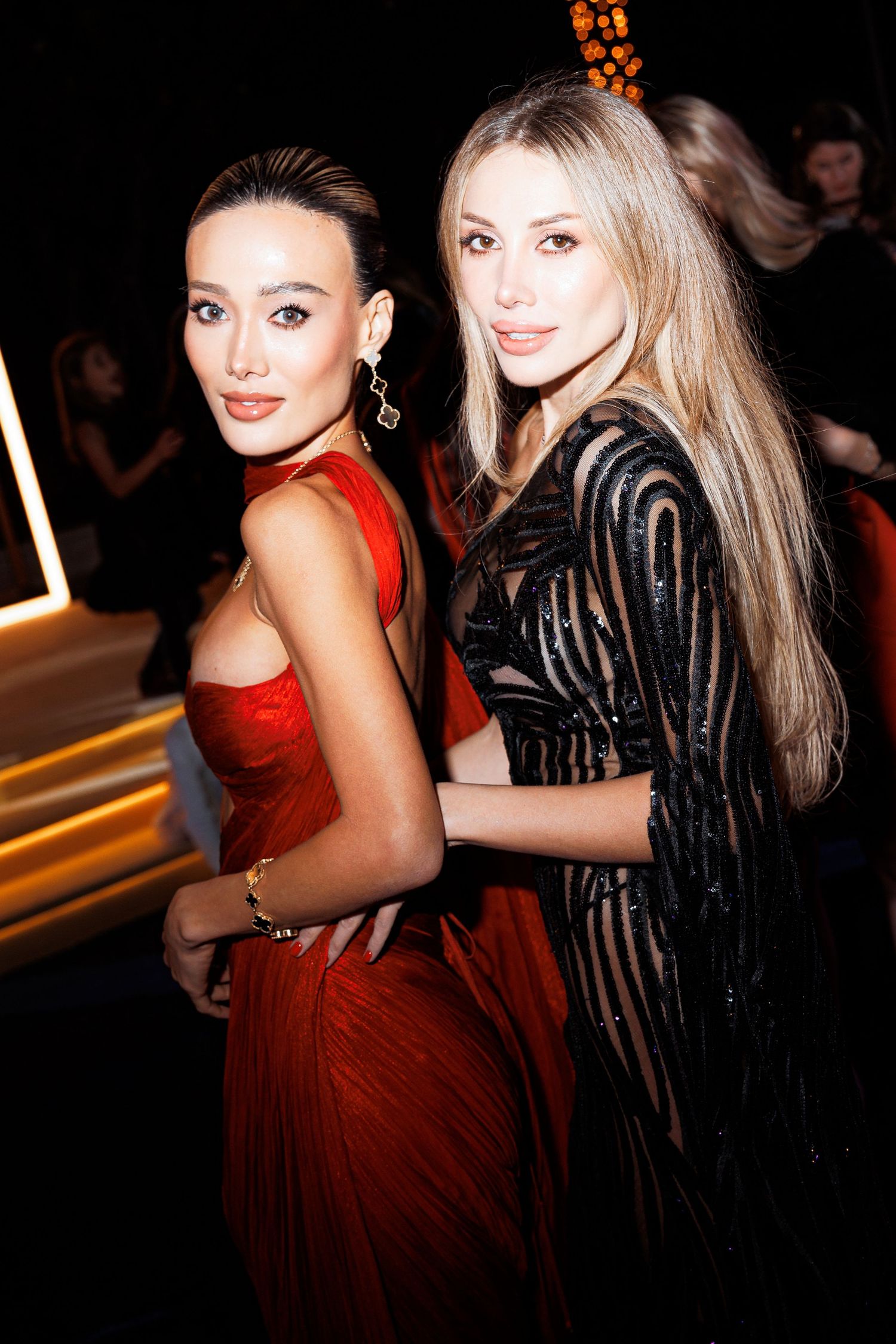Two women pose together at an event, one wearing a red dress and one in a black striped outfit.