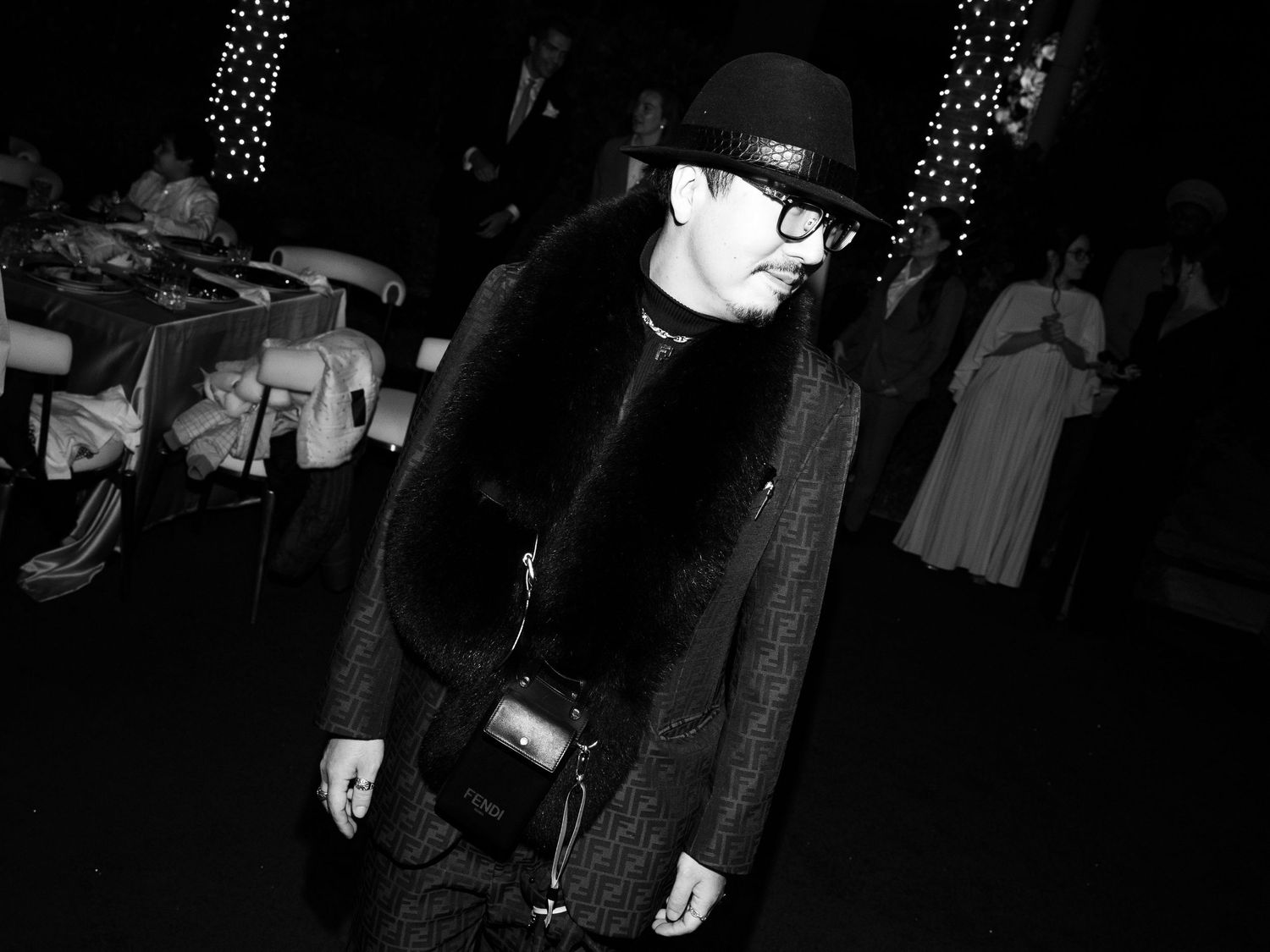 A black and white nighttime photograph showing someone in a dark coat and hat walking through a dimly lit space.