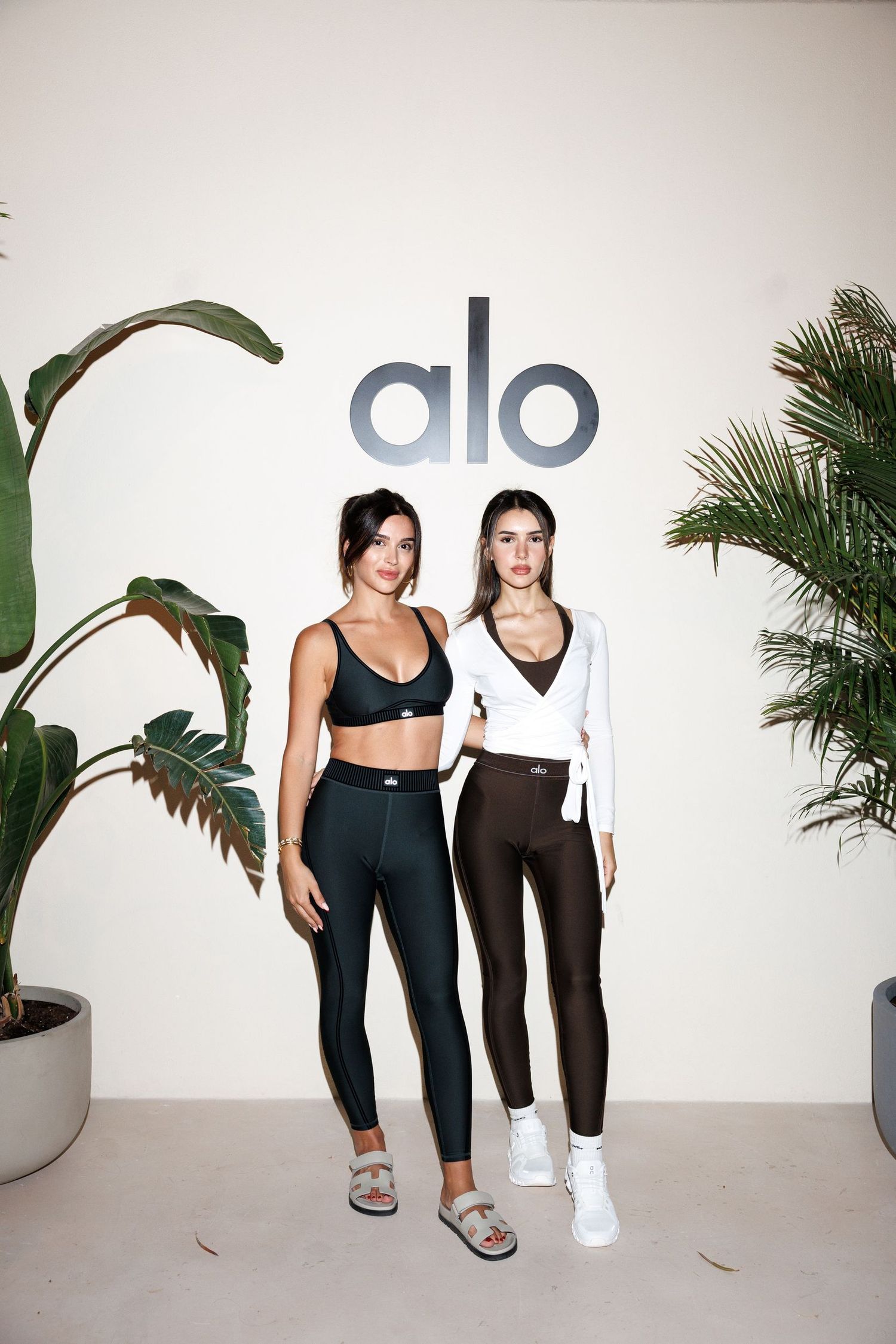 Group in athletic wear posing together against white wall with alo branding and tropical plants.