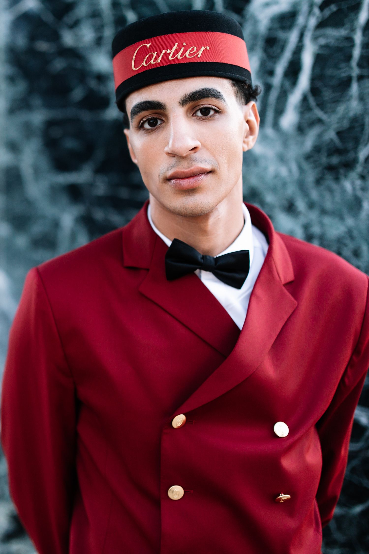 A person in a red uniform coat and black beret poses against a dark background with Cartier branding.