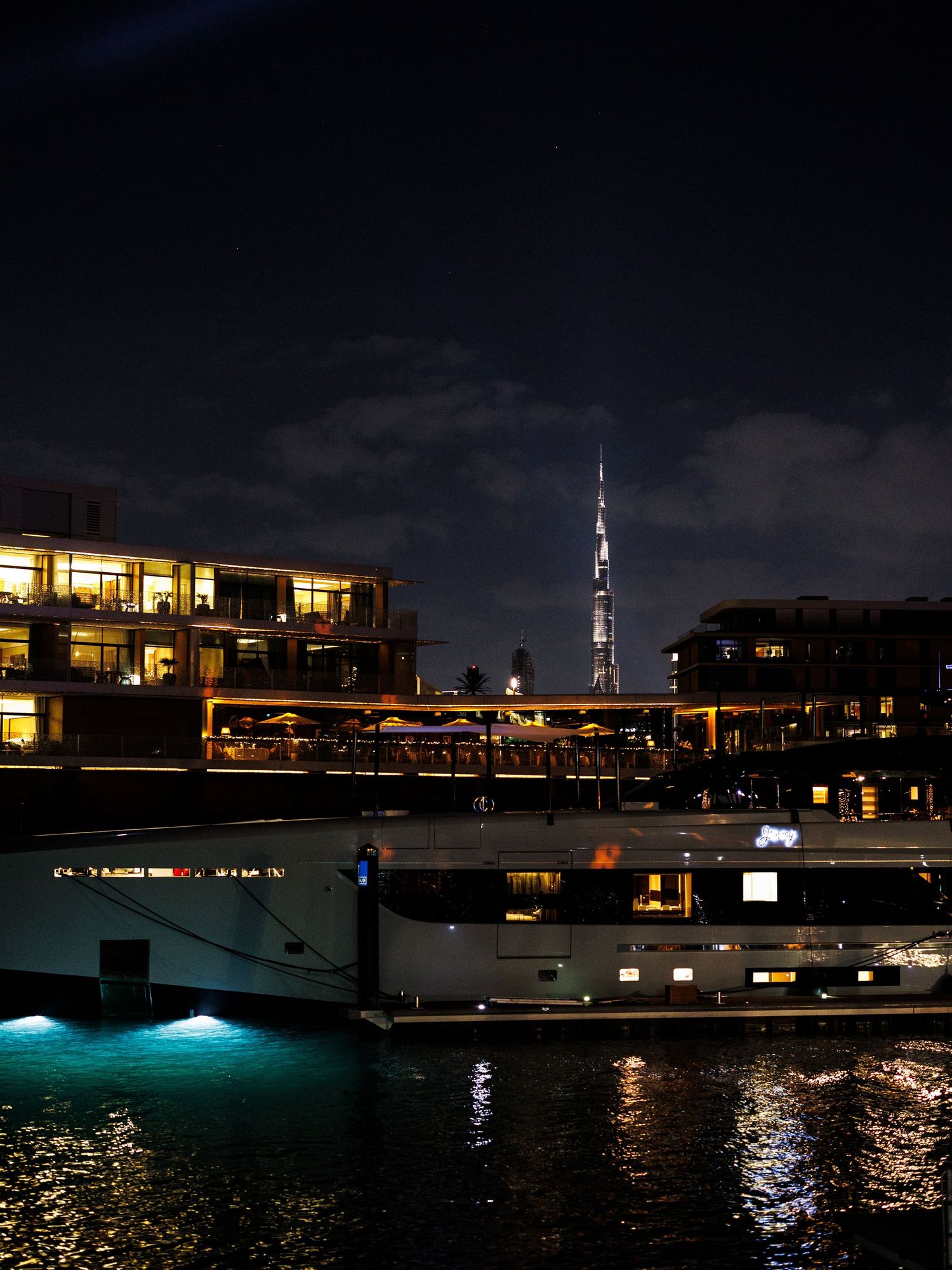 Night cityscape with illuminated buildings and boats reflecting on dark water at Dubai Marina waterfront.