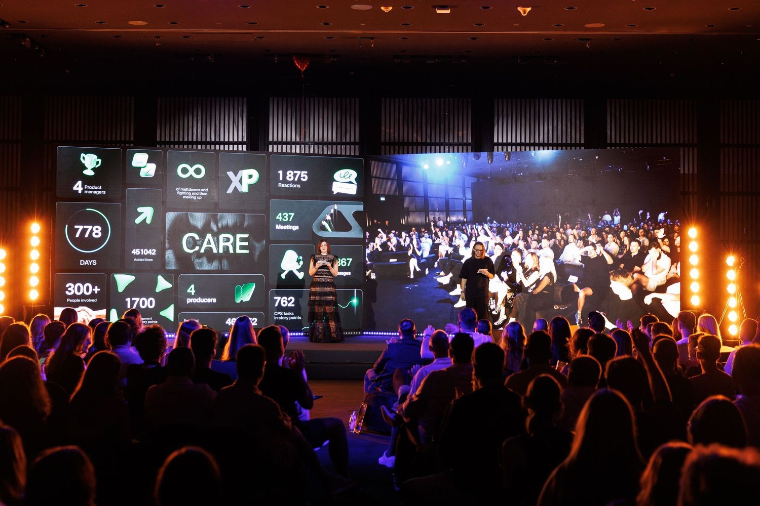 Large audience watches digital display showing logos and graphics at a corporate event with dramatic lighting.