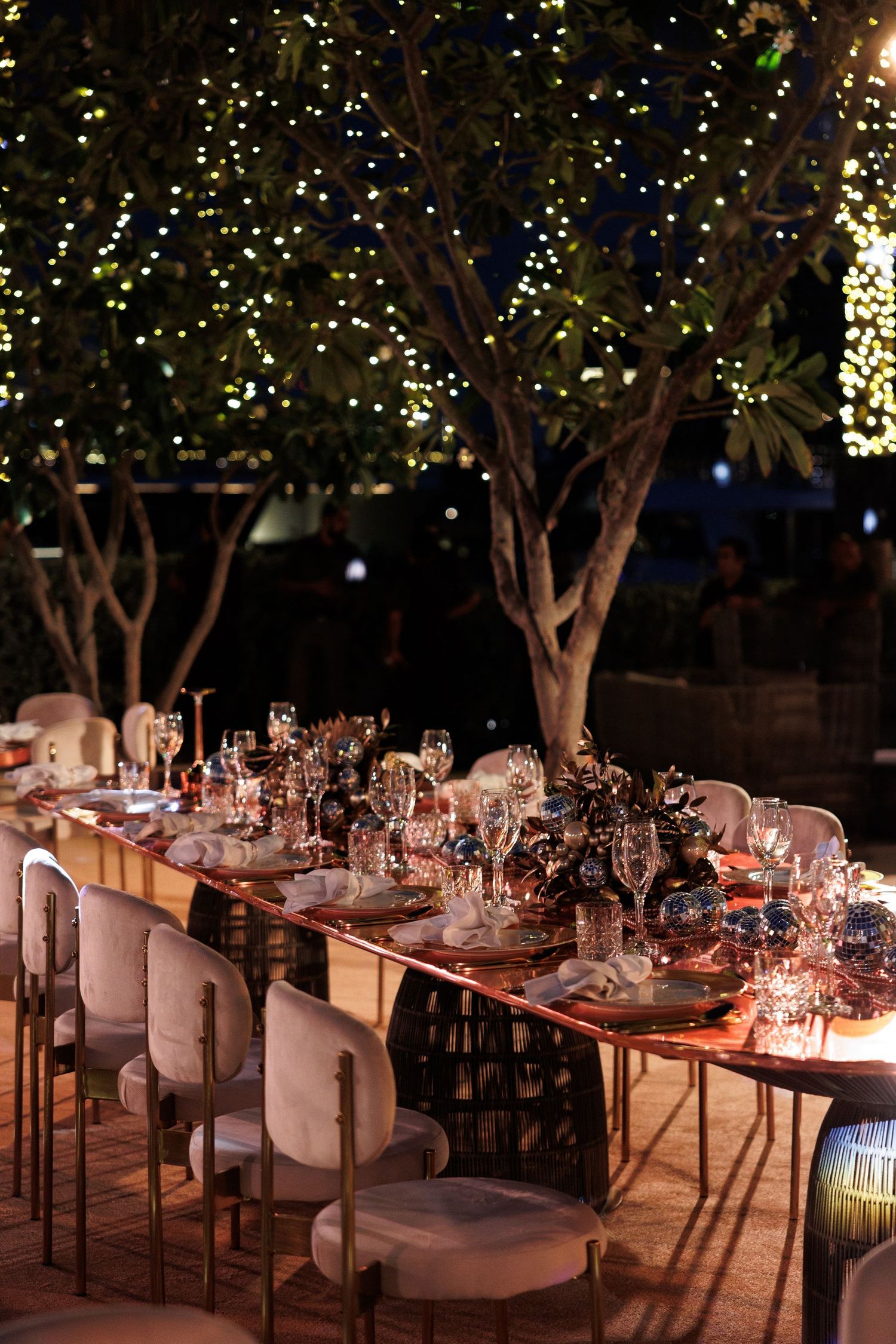 Elegant outdoor dining tables with white chairs under trees decorated with twinkling lights.