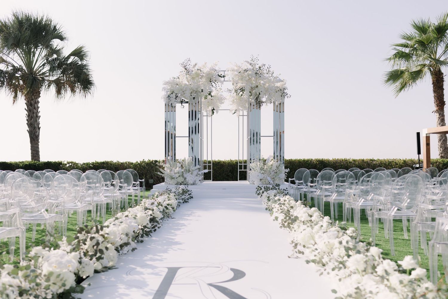 All-white wedding ceremony setup with floral arrangements and chairs under palm trees.