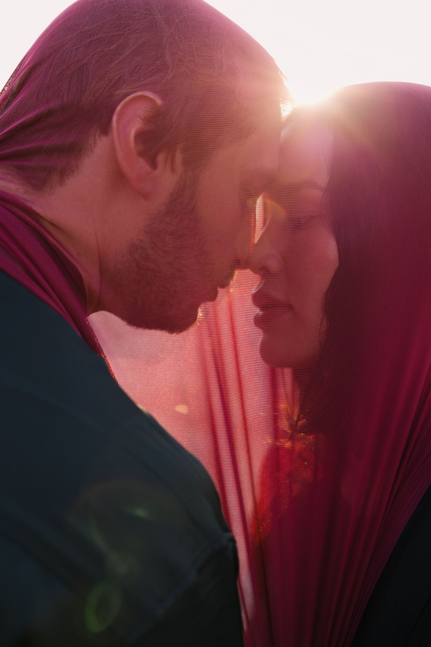 A romantic couple in silhouette sharing an intimate moment against a pink sunset with a red veil.