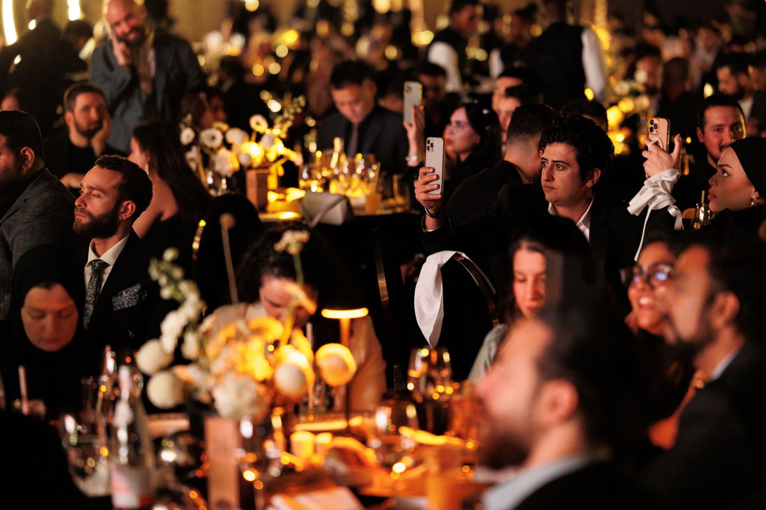 Crowded formal event with warm lighting and guests seated at elegantly decorated tables.