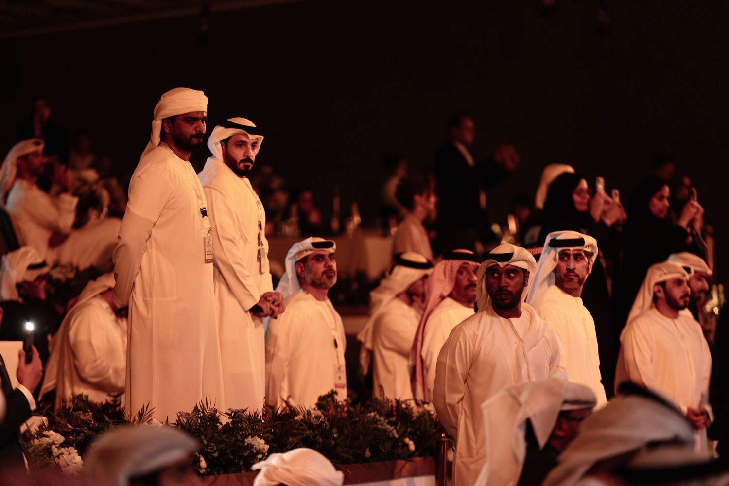 A group of people wearing white traditional clothing gather in a dimly lit space during a cultural ceremony.