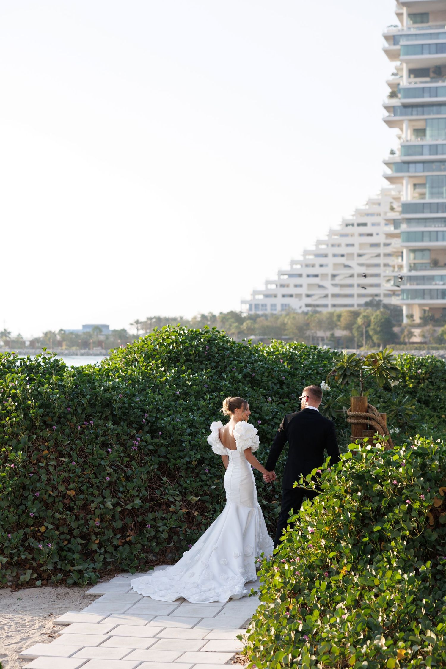 A bride in a flowing white wedding dress poses against a backdrop of modern buildings and lush greenery.
