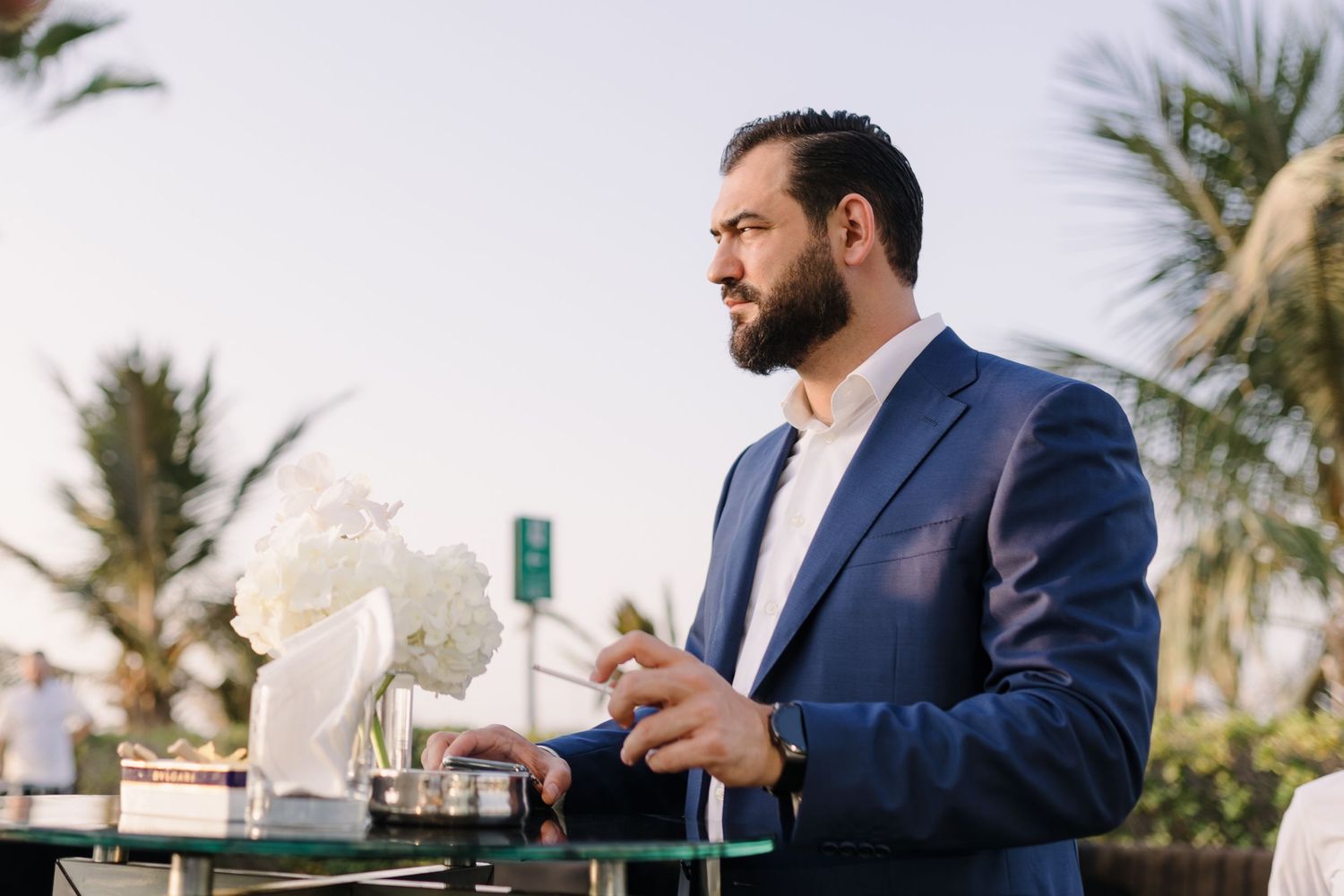 A person in a navy blazer stands outdoors near palm trees at an elegant event setup.