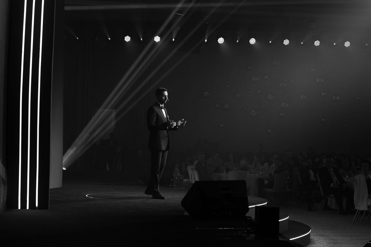 Performer stands alone on dimly lit stage with dramatic spotlight beams cutting through darkness.