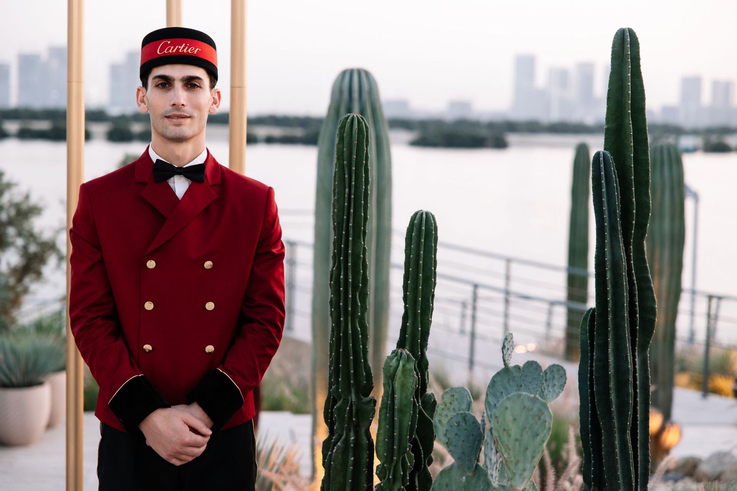 A red uniformed person stands beside tall saguaro cacti on a balcony with a city skyline in the background.