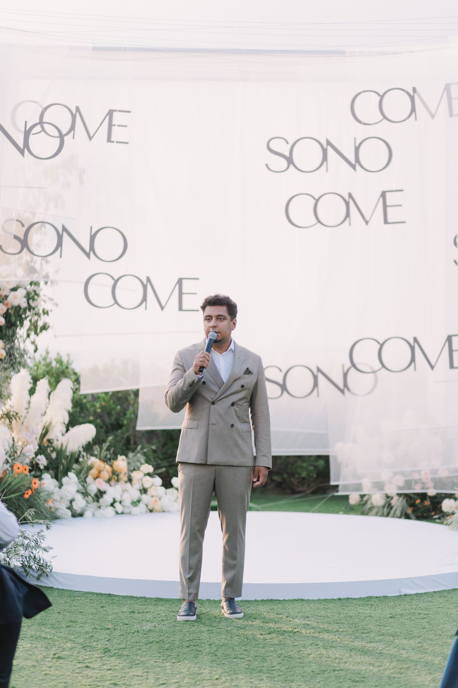 An outdoor event space with white backdrop displaying 'COME SONO' text and floral arrangements.