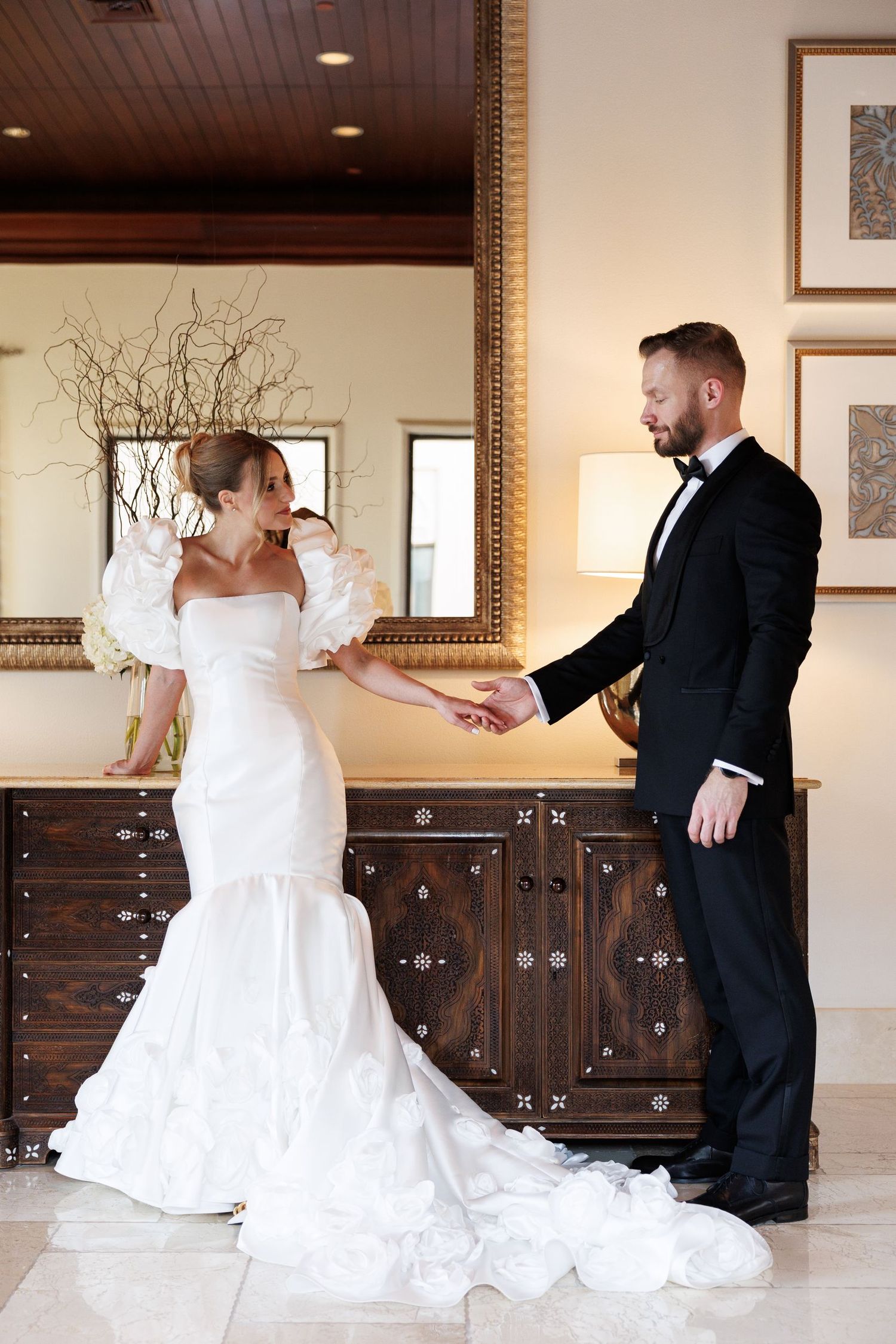 A wedding couple interacts playfully by a wooden dresser in an elegant indoor setting.