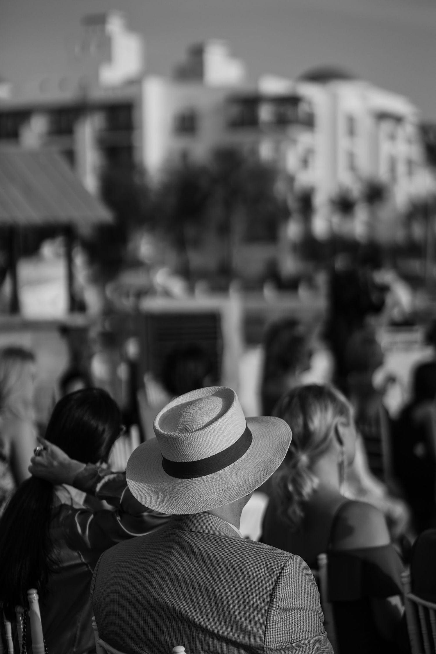 Black and white image of wedding guests wearing formal hats at an outdoor event.