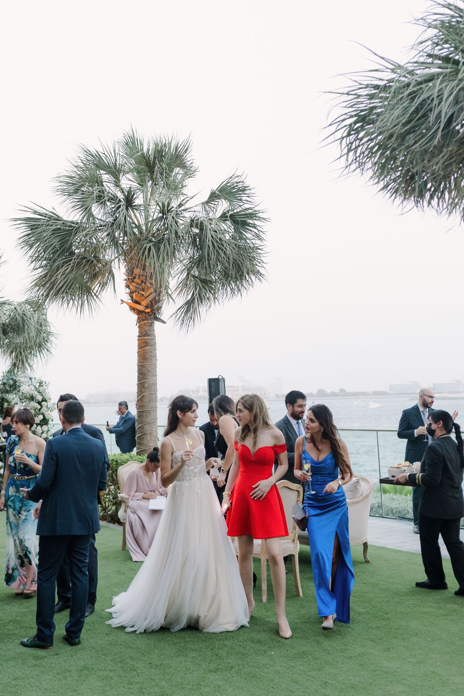 Wedding guests gather on a lawn with palm trees and ocean views during an outdoor reception.