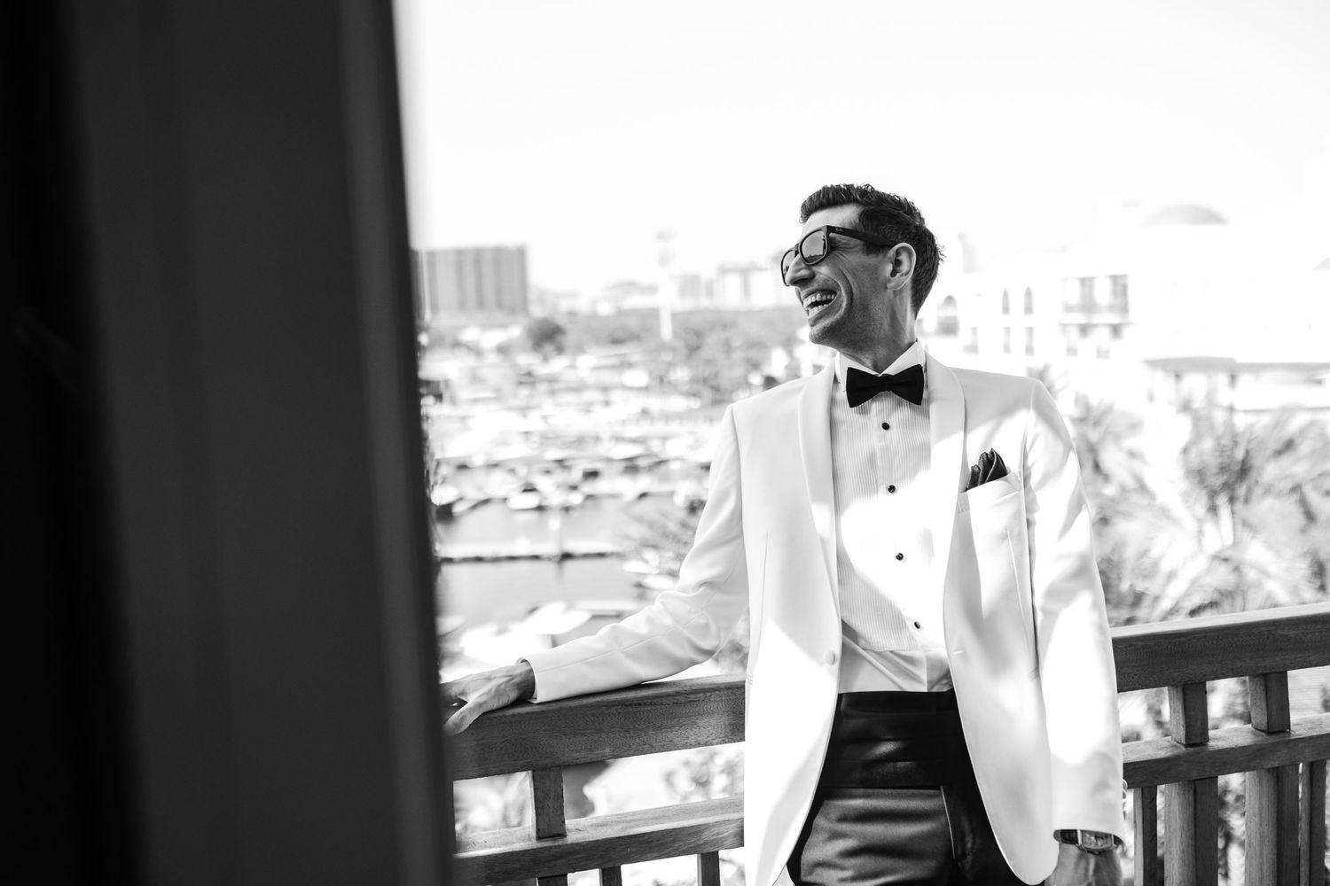 Black and white portrait of groom in white tuxedo leaning on balcony railing.