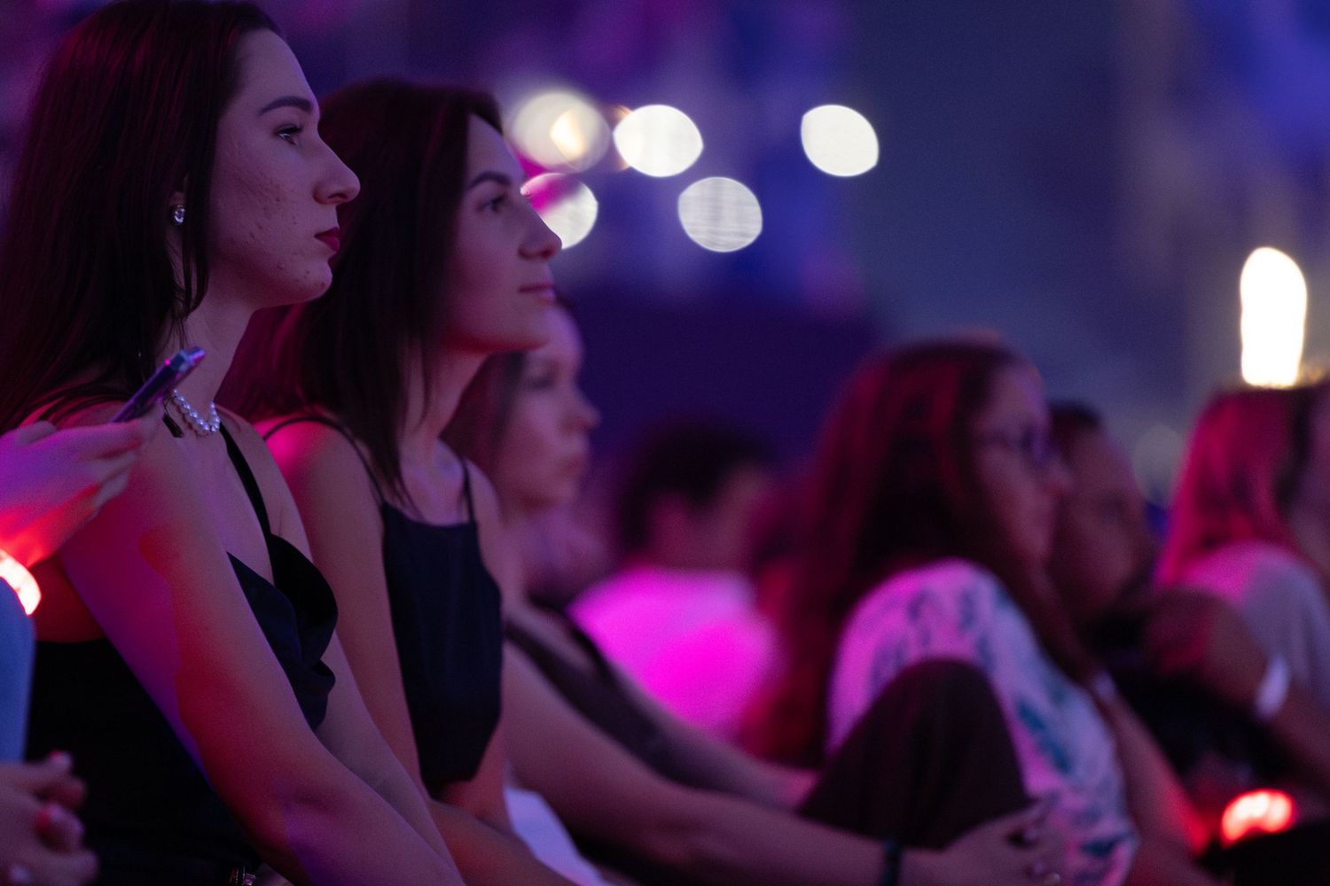 Audience members watch intently in a darkened venue with glowing bokeh lights in the background.