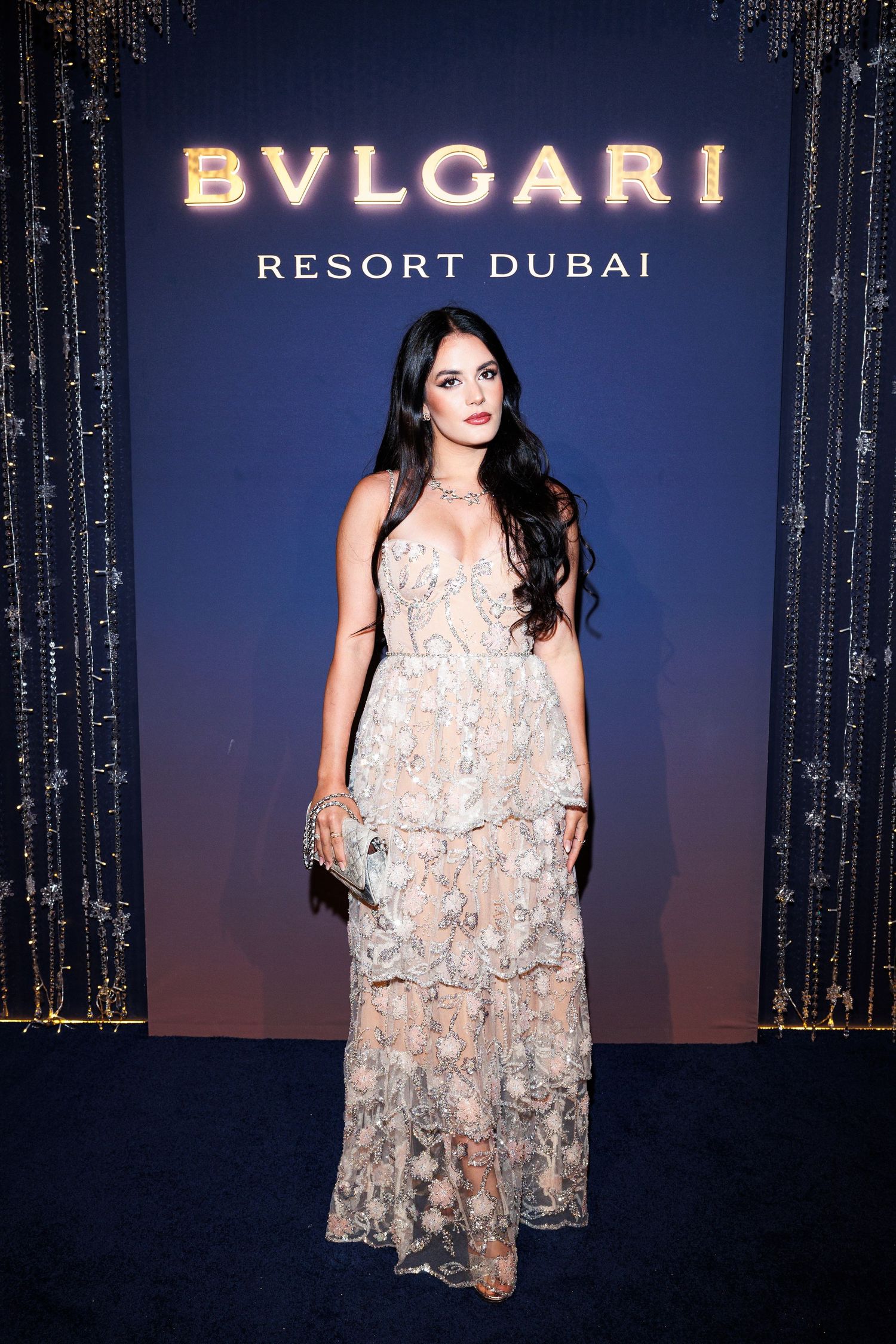 A person wearing a sheer floral embellished dress poses at a Bulgari Resort Dubai event against a dark backdrop.