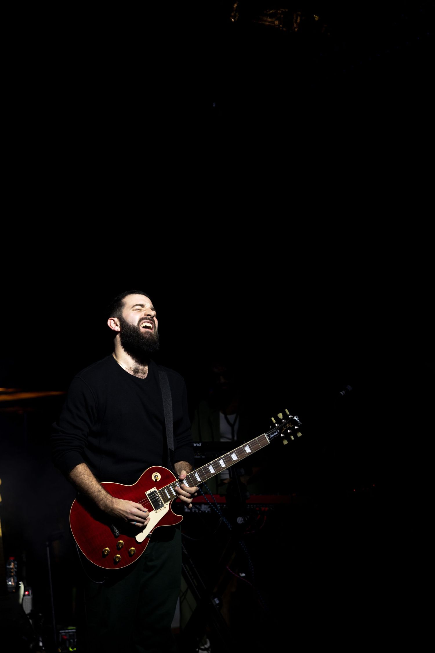 A guitarist in black clothing performs on stage with a red Gibson Les Paul guitar in dramatic lighting.