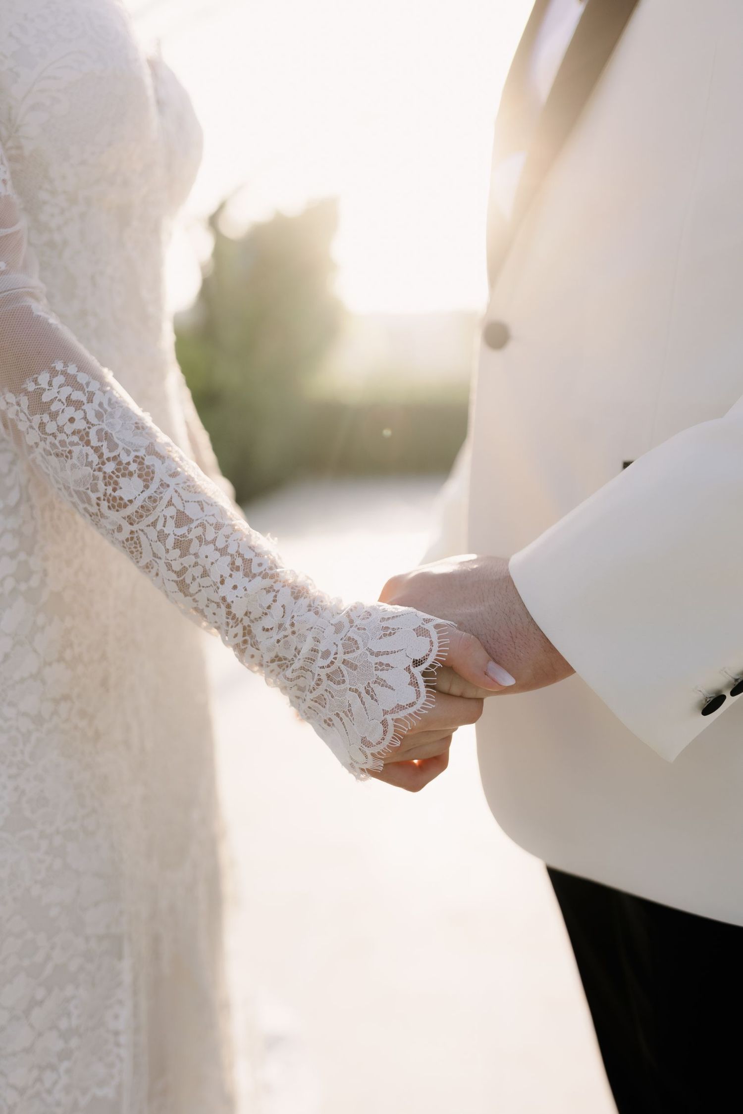 Close-up of delicate lace details on a white wedding dress with hands clasped.