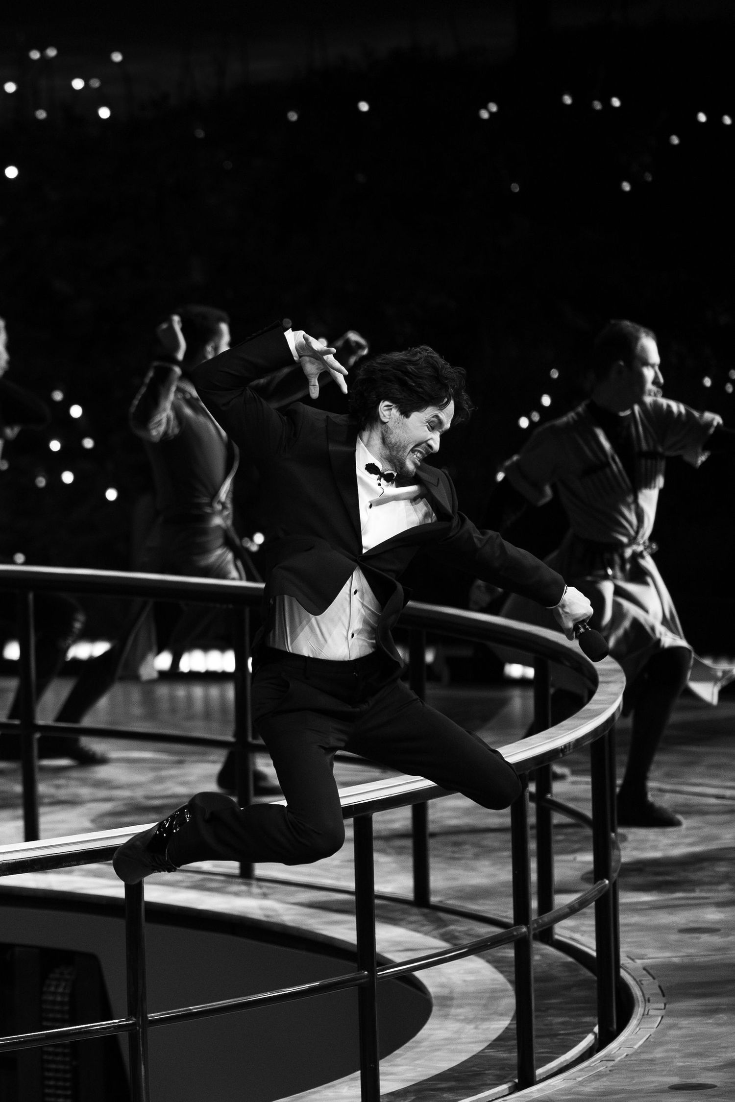 Performers in formal attire leap over barriers in a synchronized dance routine.