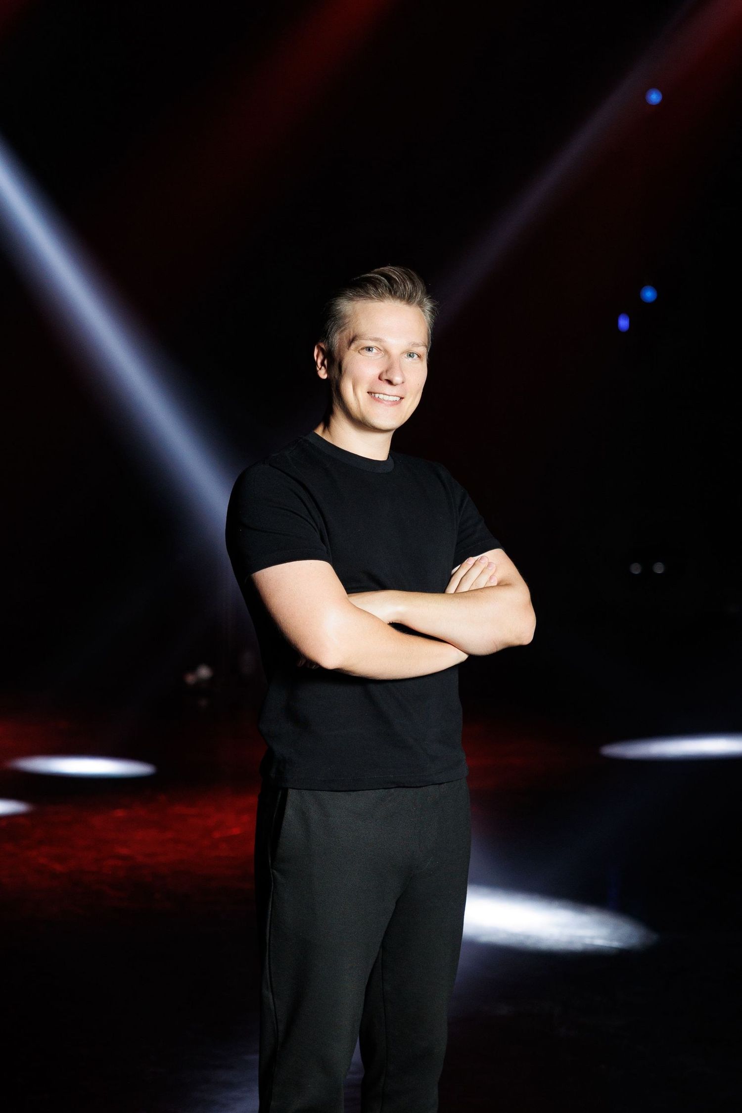 Performer in black clothing posing on dark stage with spotlights.