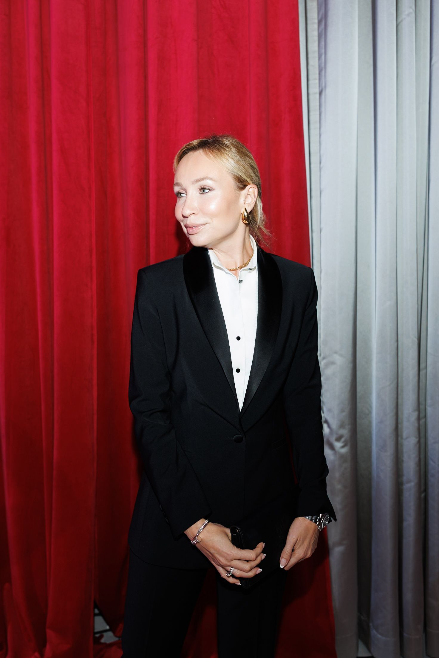 A person wearing a classic black tuxedo poses in front of red and gray curtains.