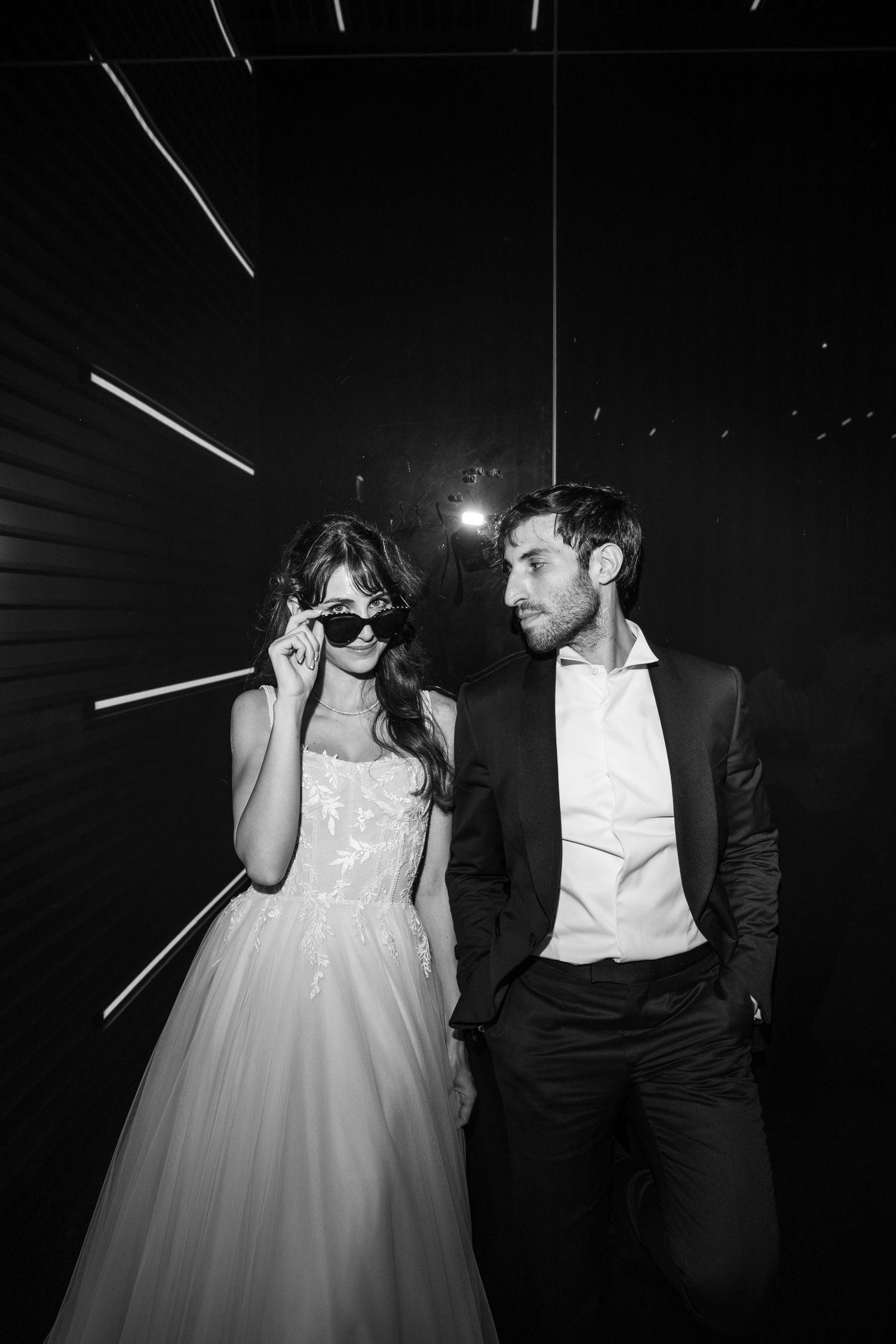 A stylish couple poses against a dark background with dramatic lighting and the bride wearing sunglasses.