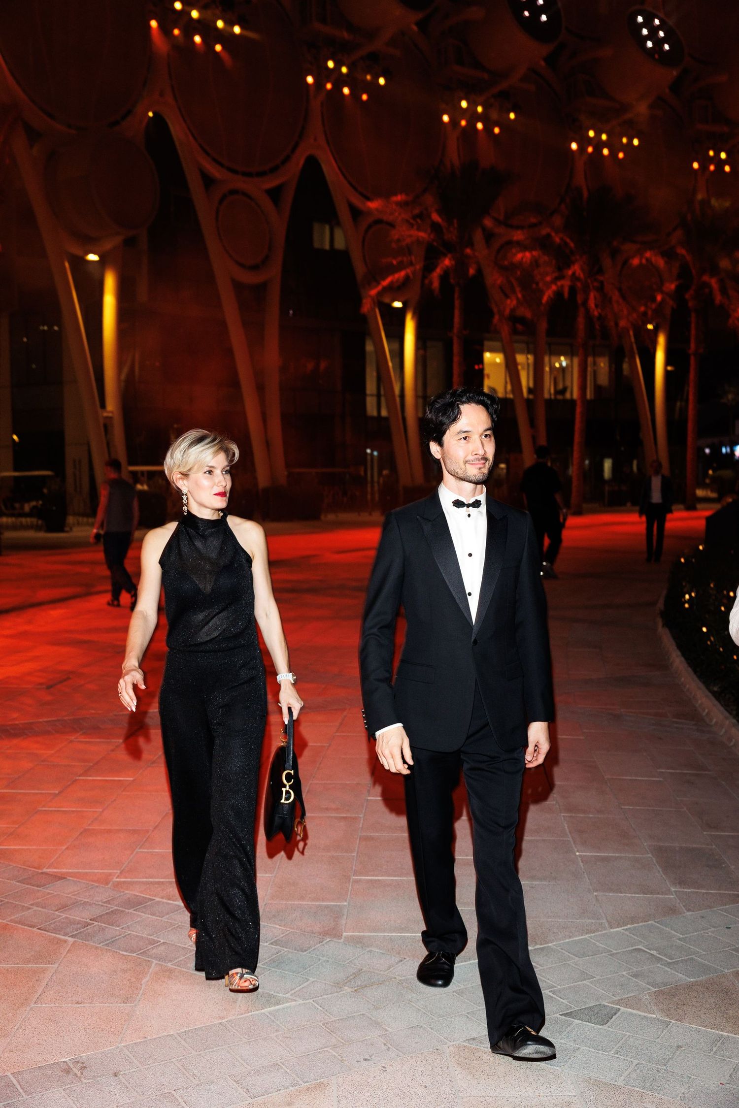People in formal attire walk across a red-lit stone patio during an evening event.