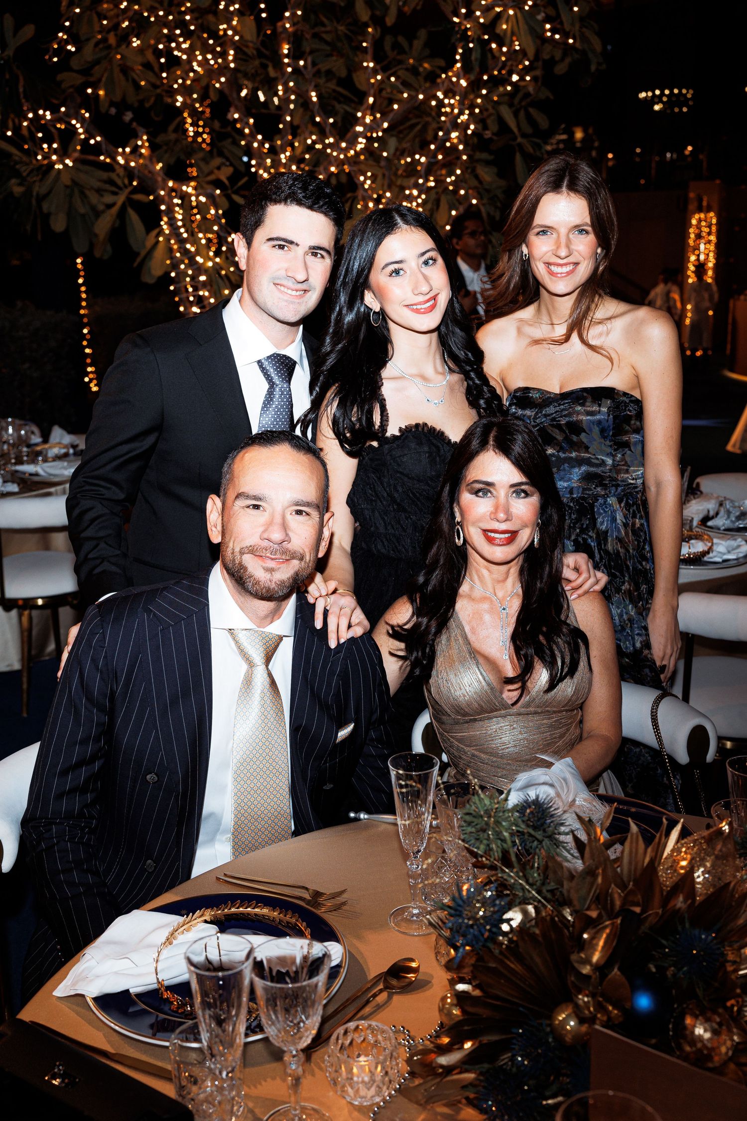 Elegant party guests gather around a decorated table at an evening event with twinkling lights in the background.