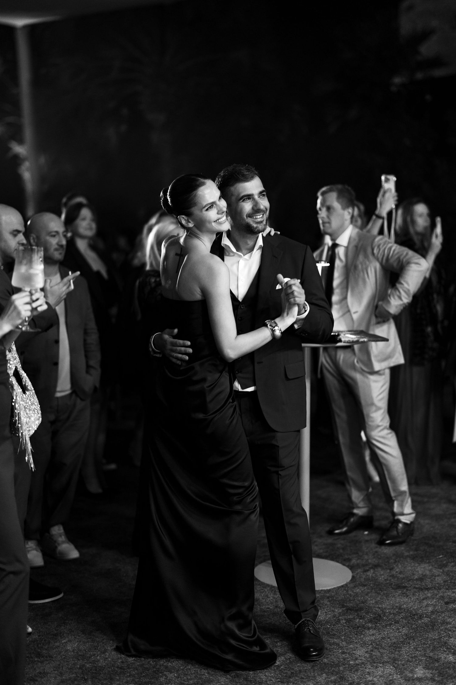 A couple shares an intimate slow dance at an elegant evening event while other guests mingle in the background.