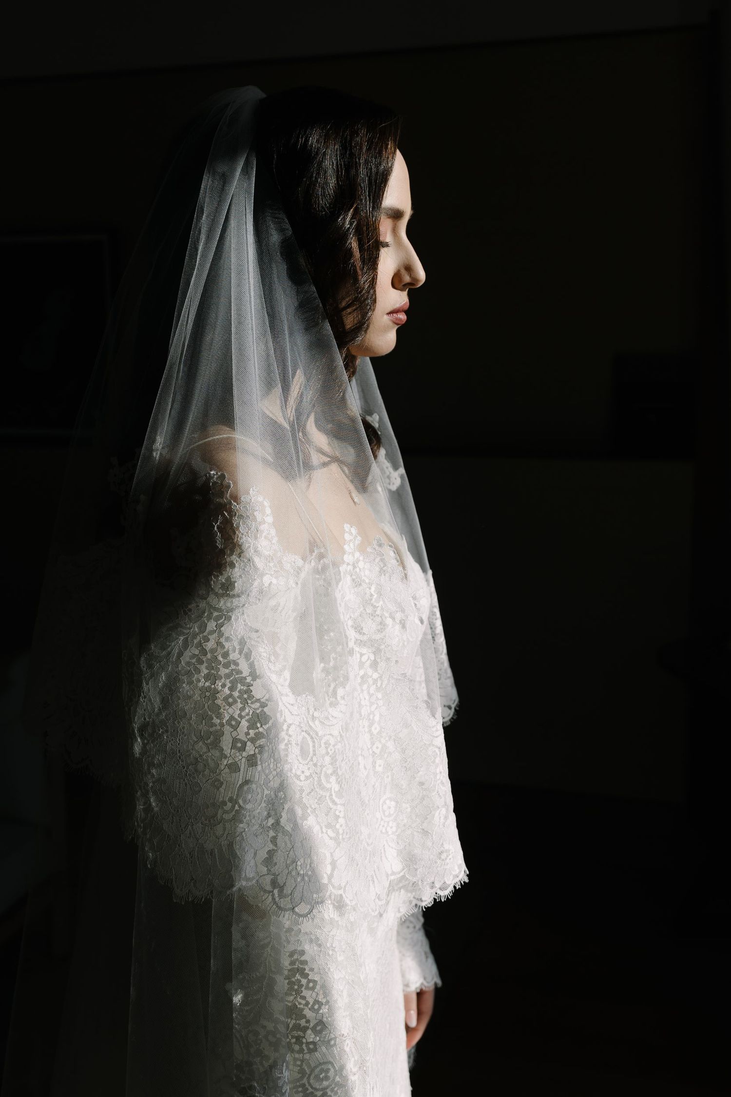 A bride in a delicate lace wedding gown and veil stands against a dark background in dramatic lighting.