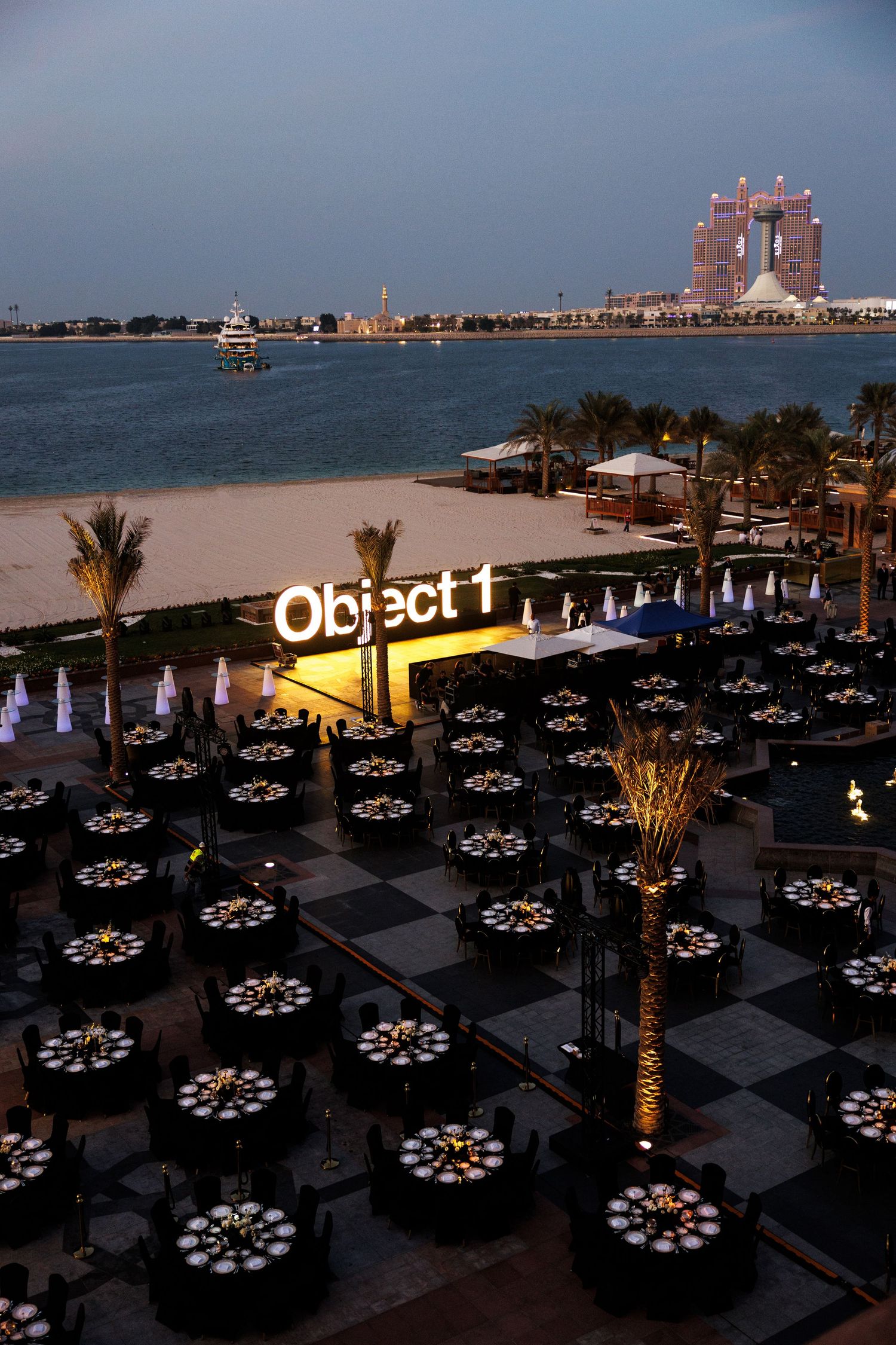 Outdoor event venue with circular dining tables arranged near a beach with palm trees and a modern building in the background.