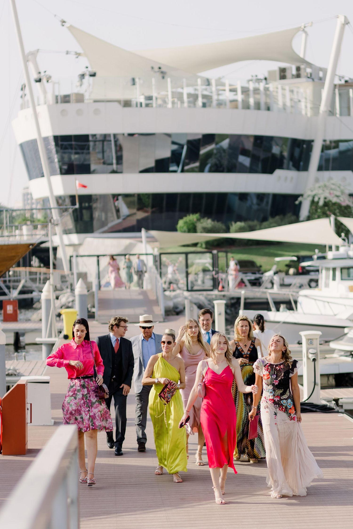 Wedding party in colorful dresses walking along a marina dock with yachts in the background.