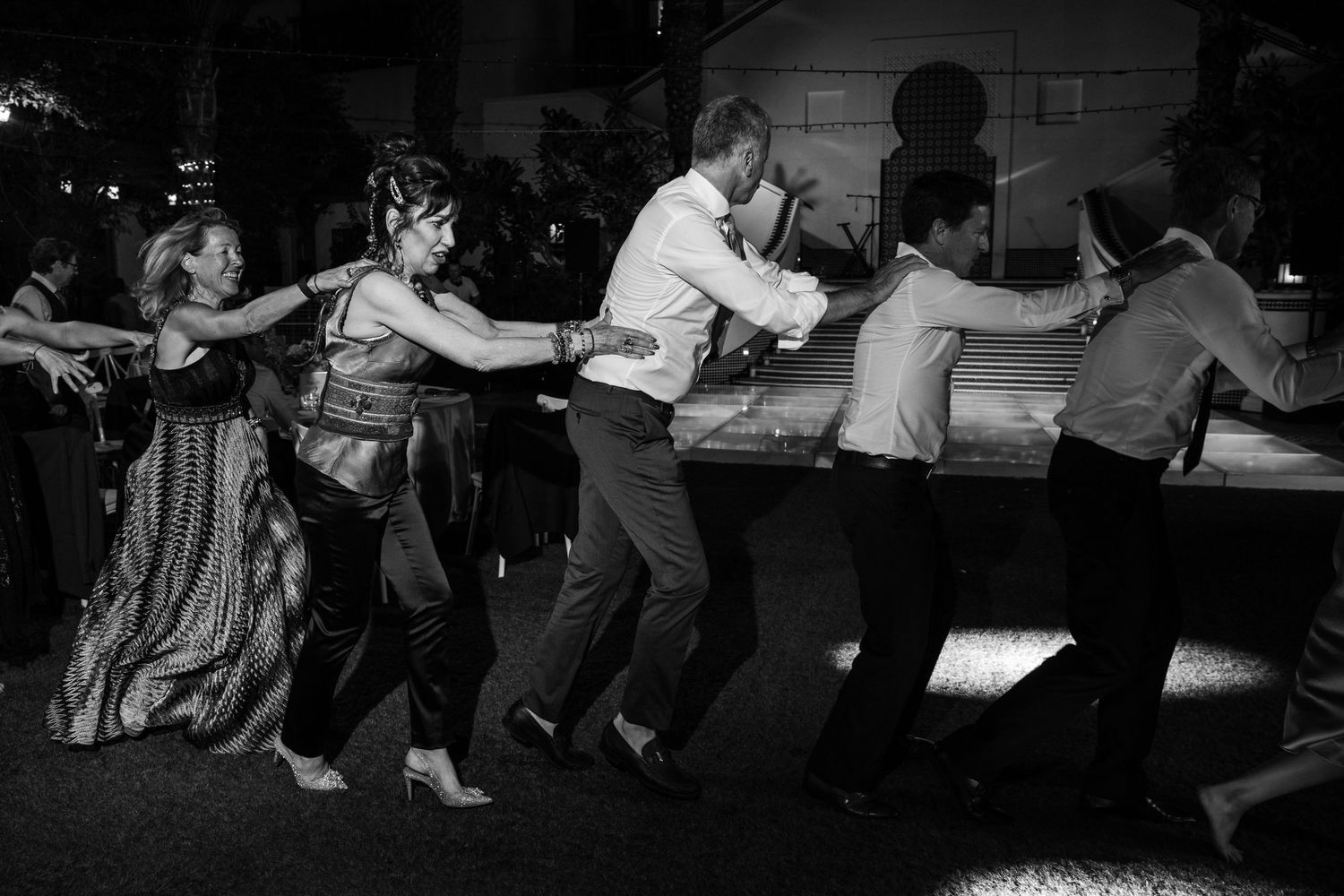 Black and white photo of people dancing in a line at a wedding reception.