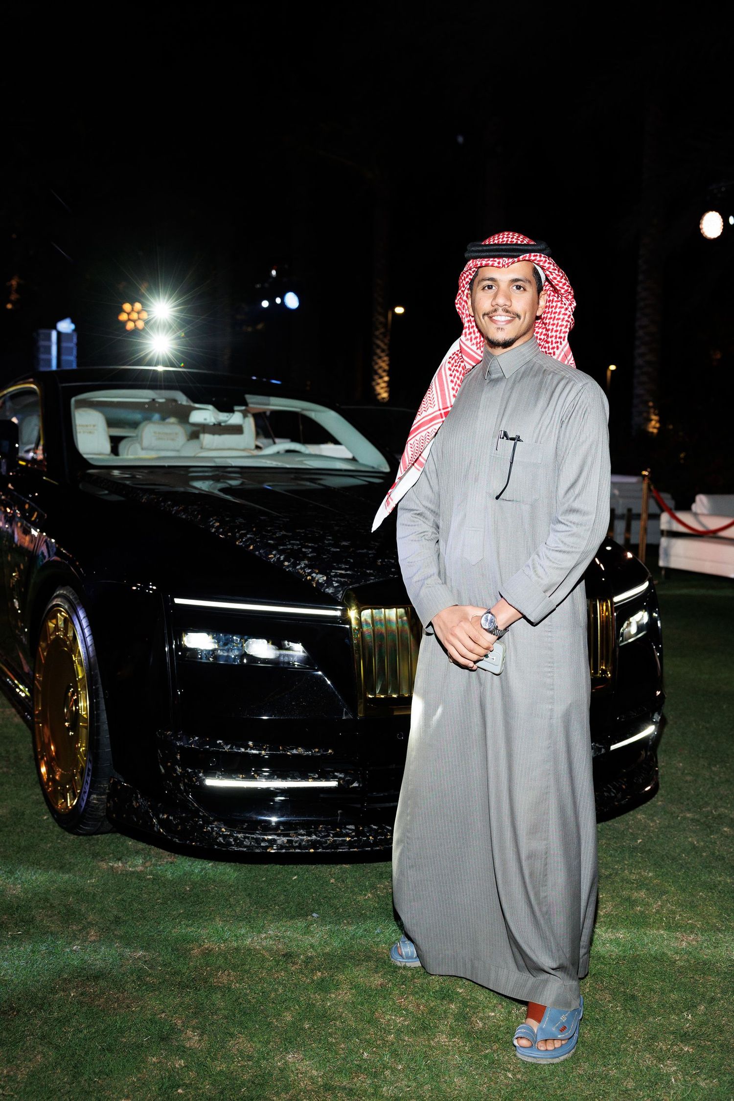 Person in traditional Middle Eastern dress standing next to a luxury black car at night.